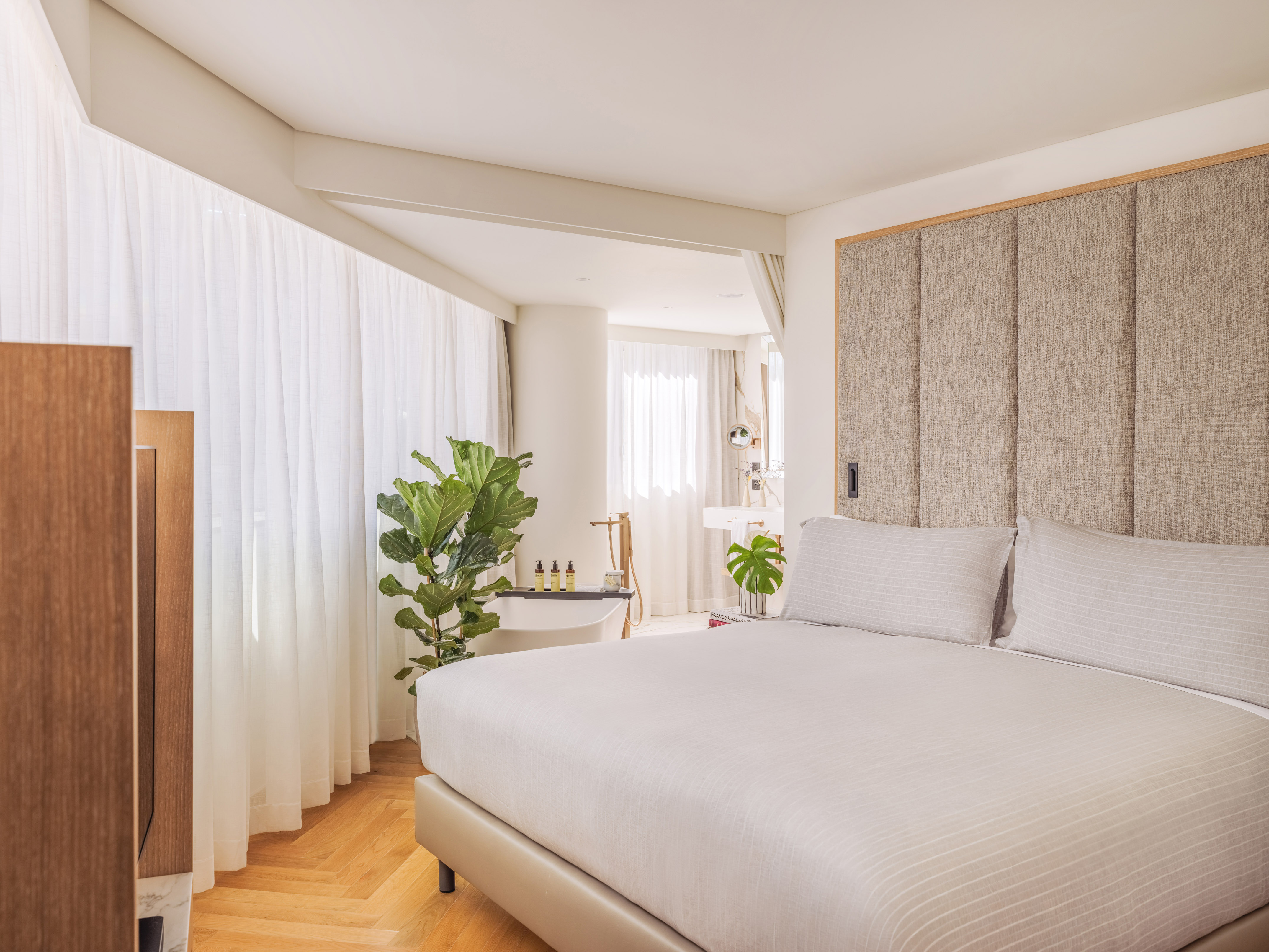 Bright hotel bedroom with king bed, fabric headboard, plant, and bathtub.