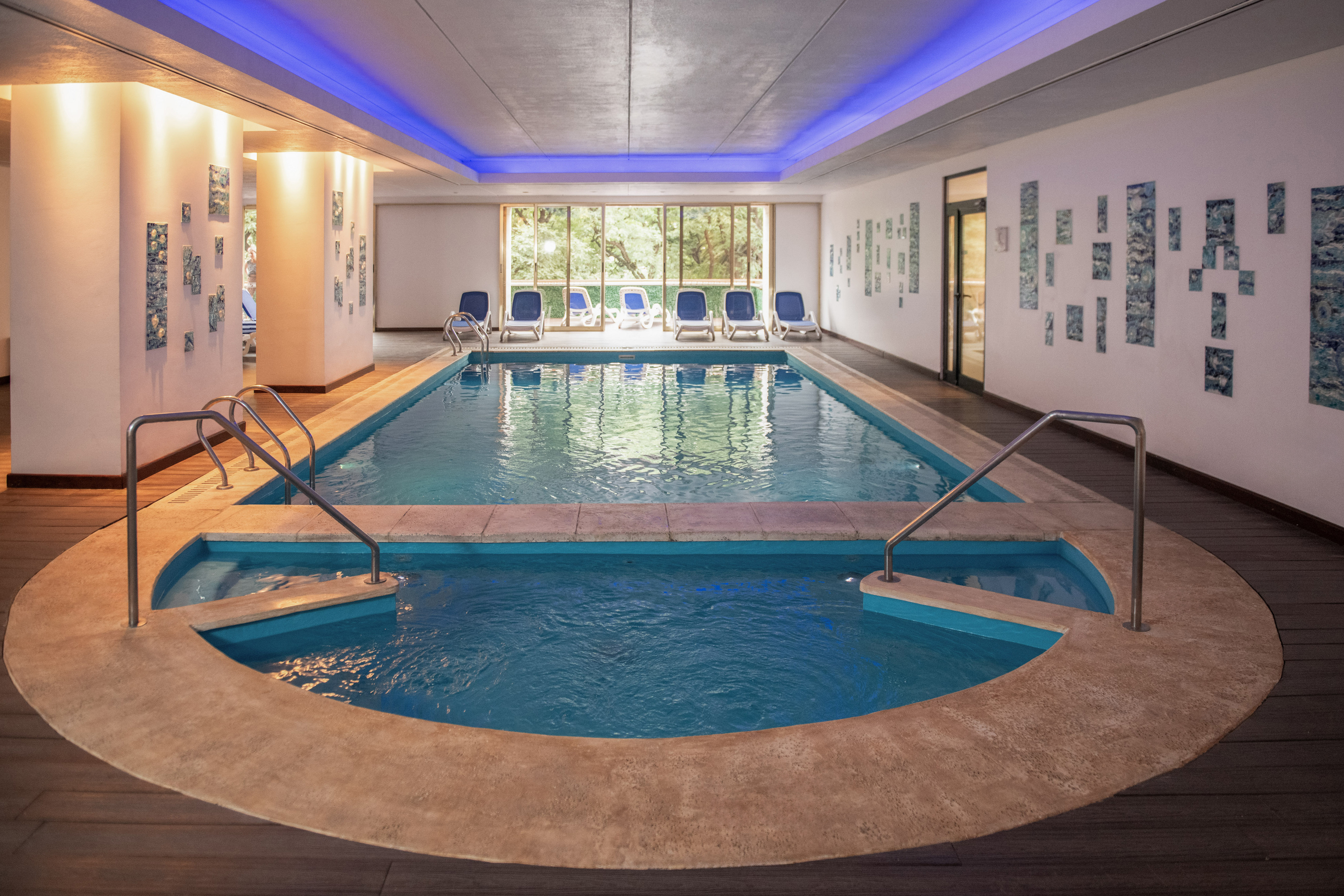 Modern indoor swimming pool with blue LED lighting, jacuzzi, and forest view.