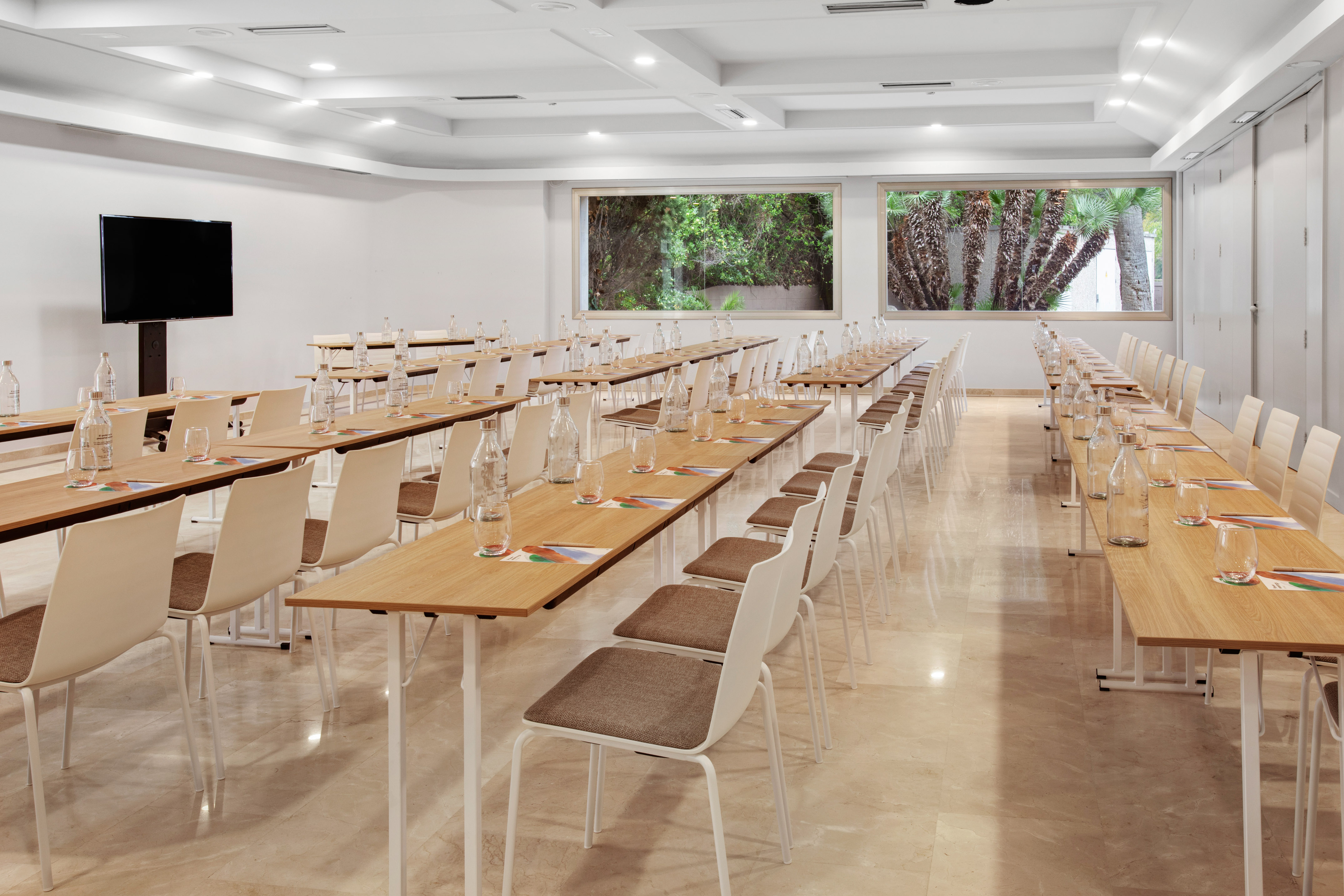 Modern conference room setup with tables, chairs, TV, and green views from windows.