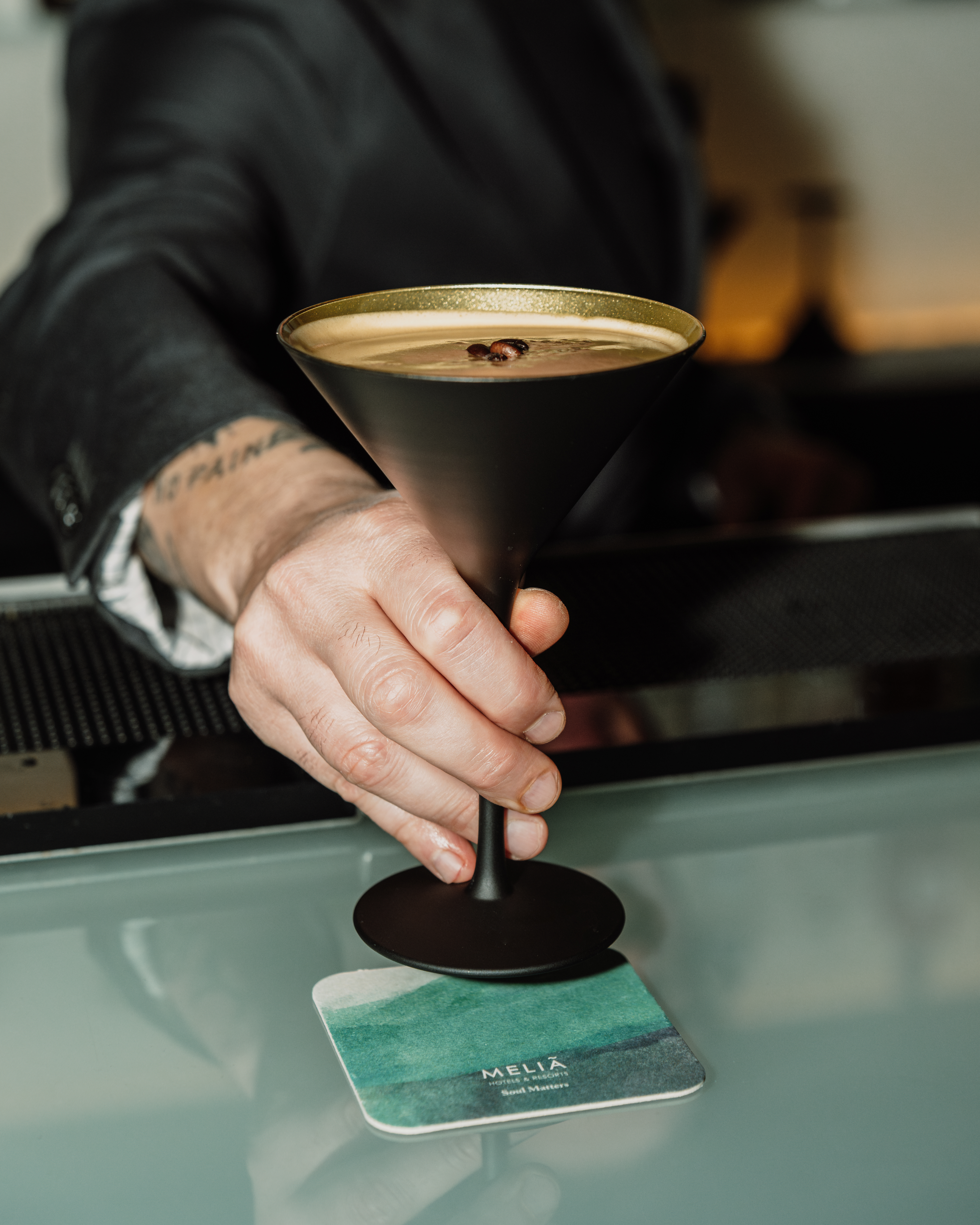 Espresso martini in black glass with coffee beans, held above Meliá coaster.