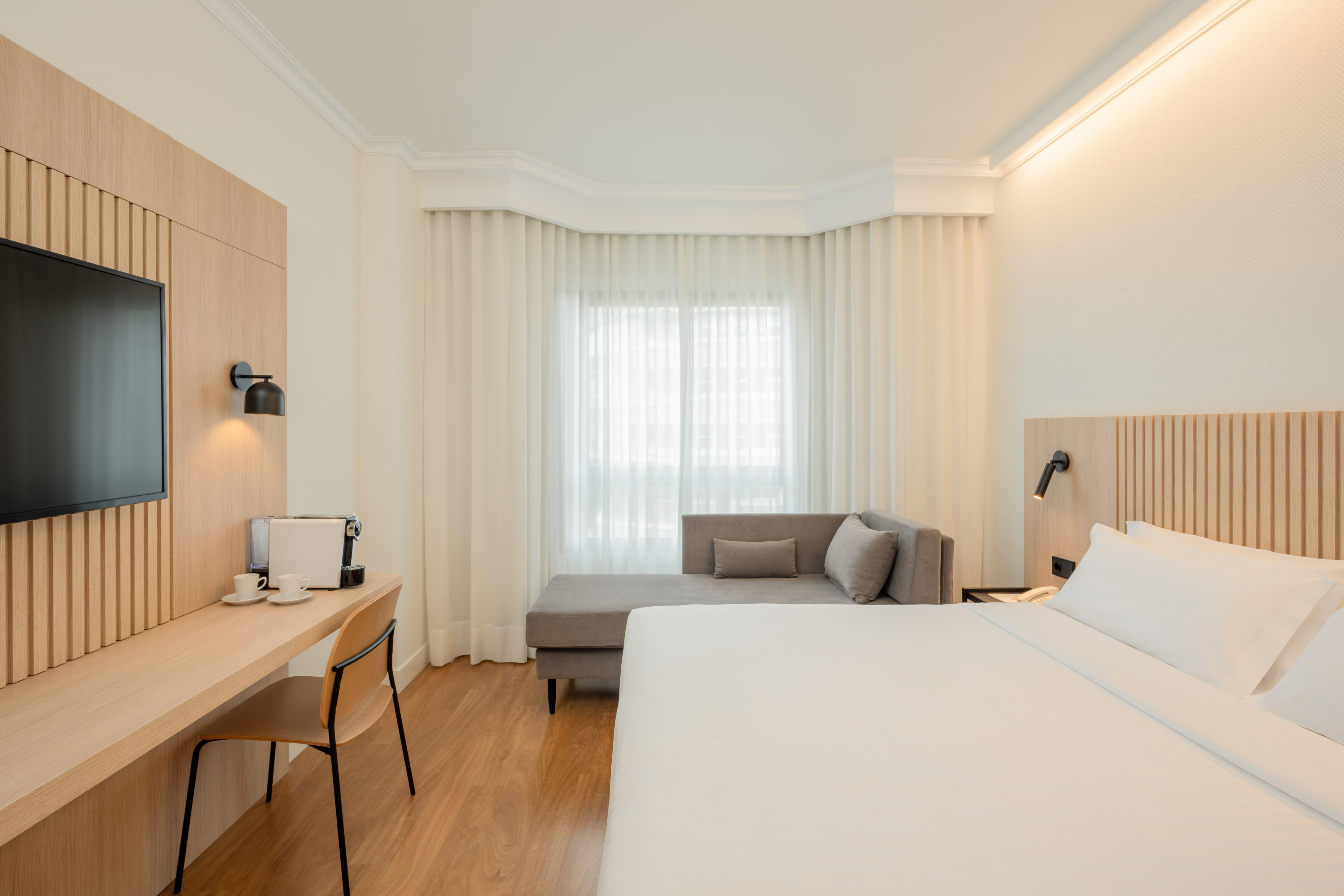 Modern hotel room with king bed, wooden work desk, TV, and grey lounge sofa.