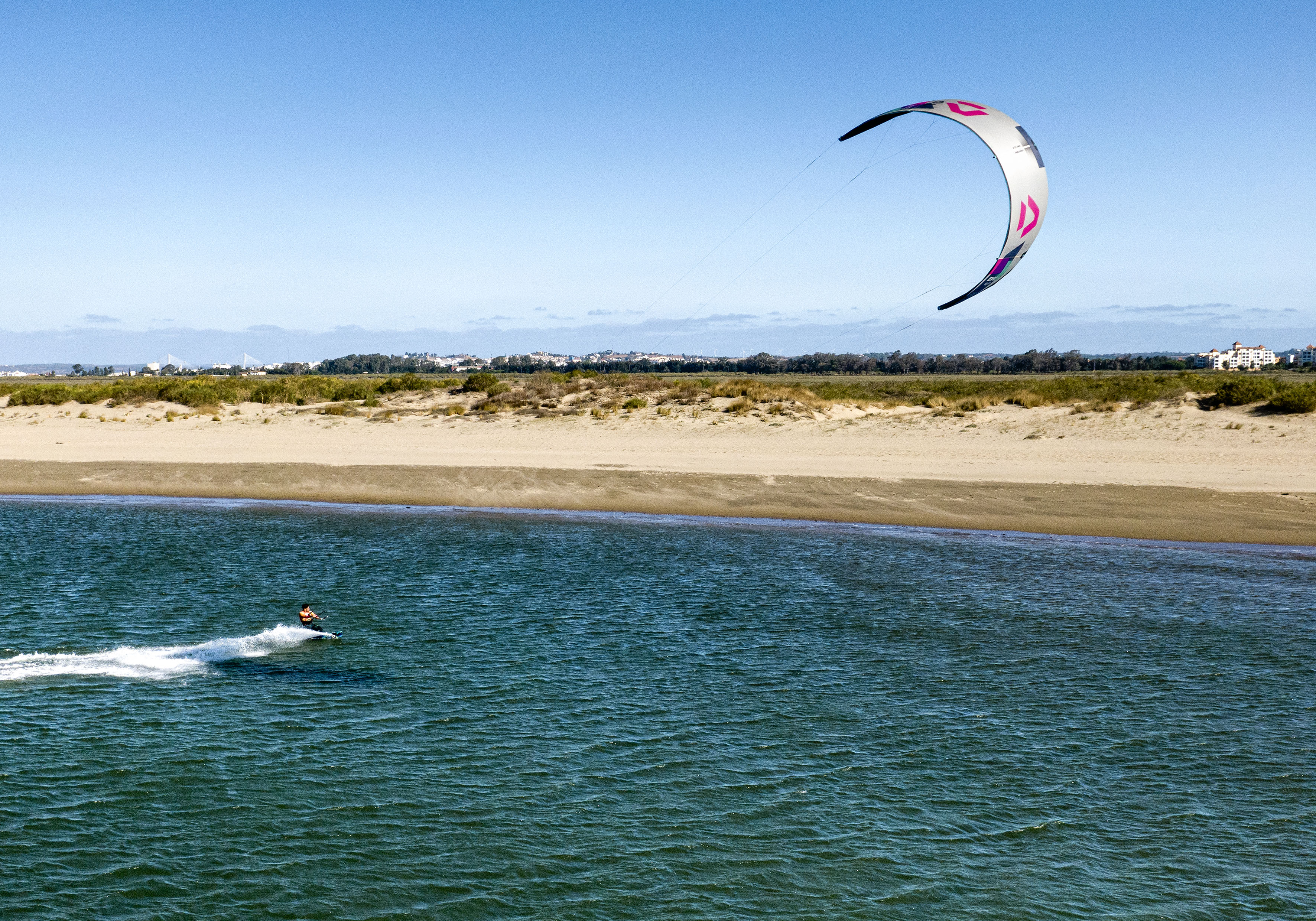 Kitesurfer on blue water with a white and pink kite, sandy beach.