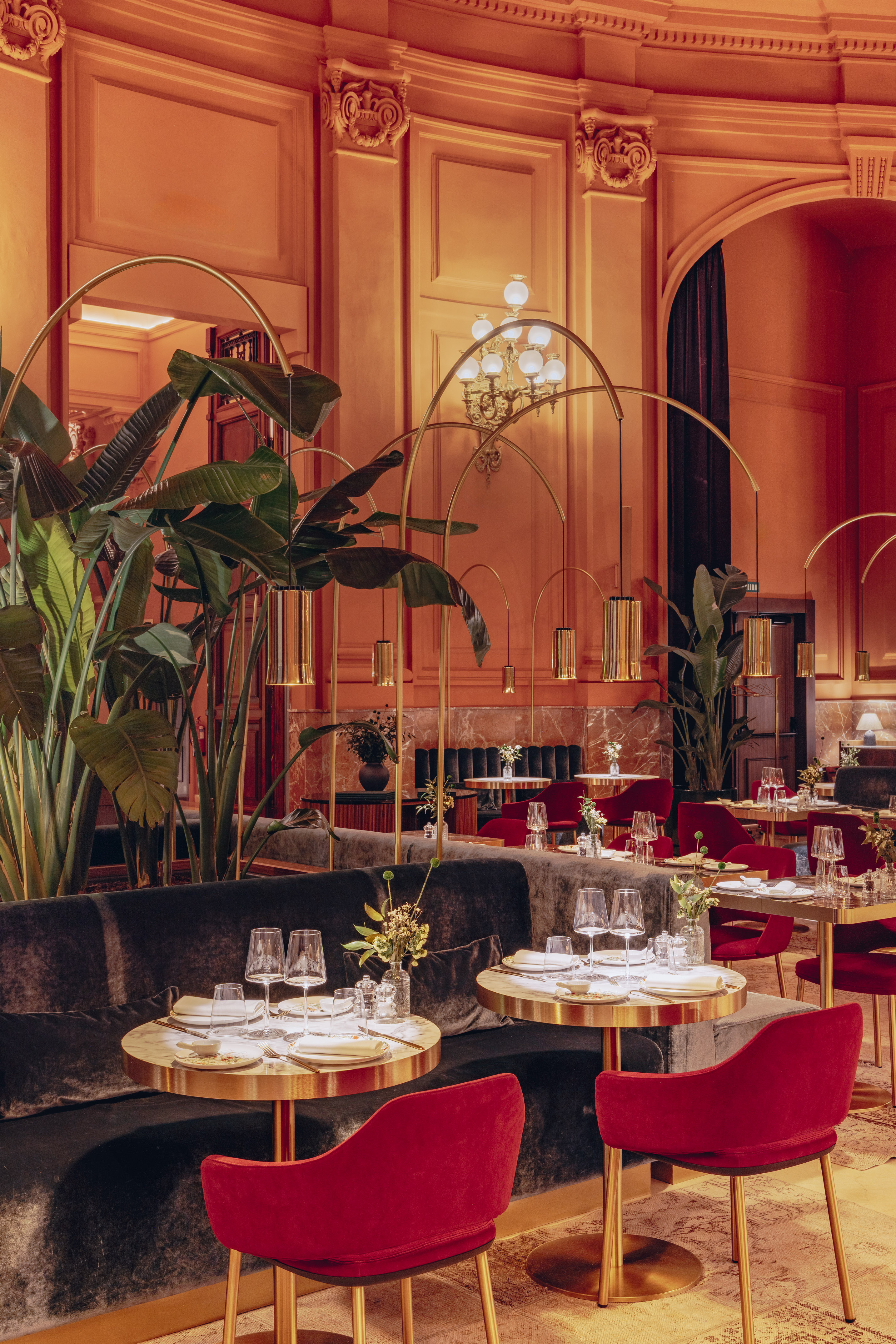 Luxury restaurant interior: red velvet chairs, gold tables, tropical plants, orange walls.