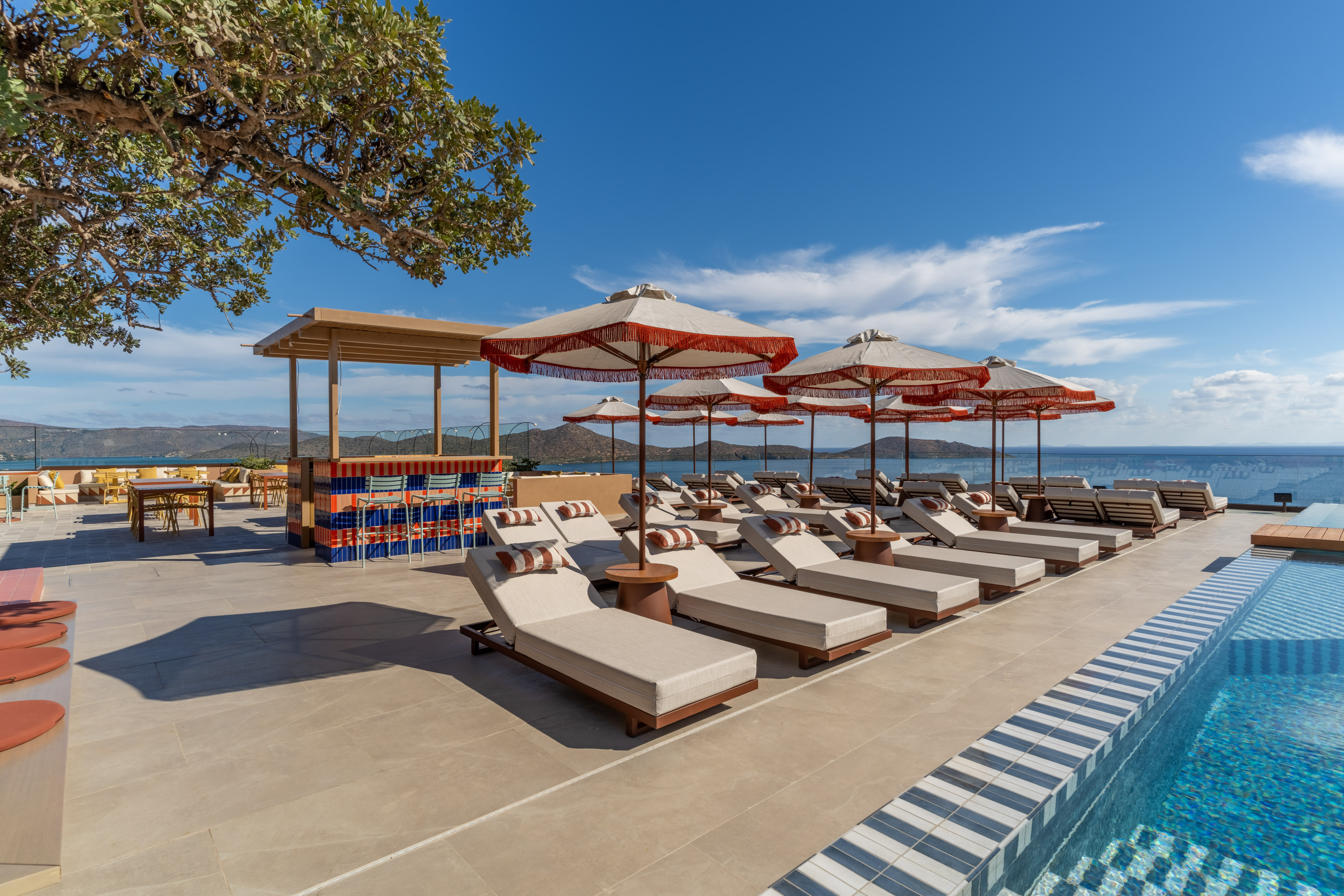 Luxury poolside bar with lounge chairs, red umbrellas, pool, and ocean view.