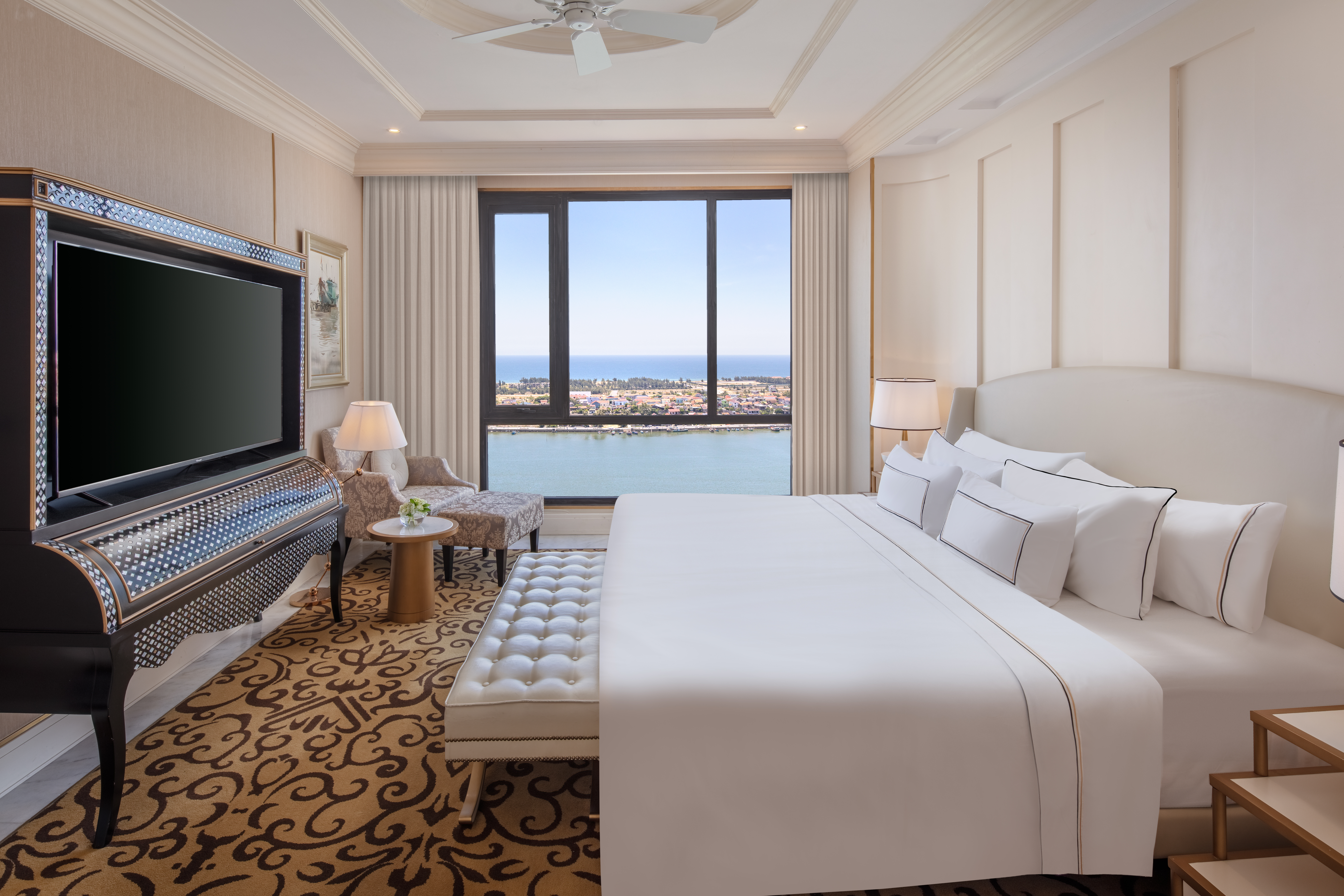 Elegant hotel bedroom suite with waterfront view, king bed, and ornate TV.