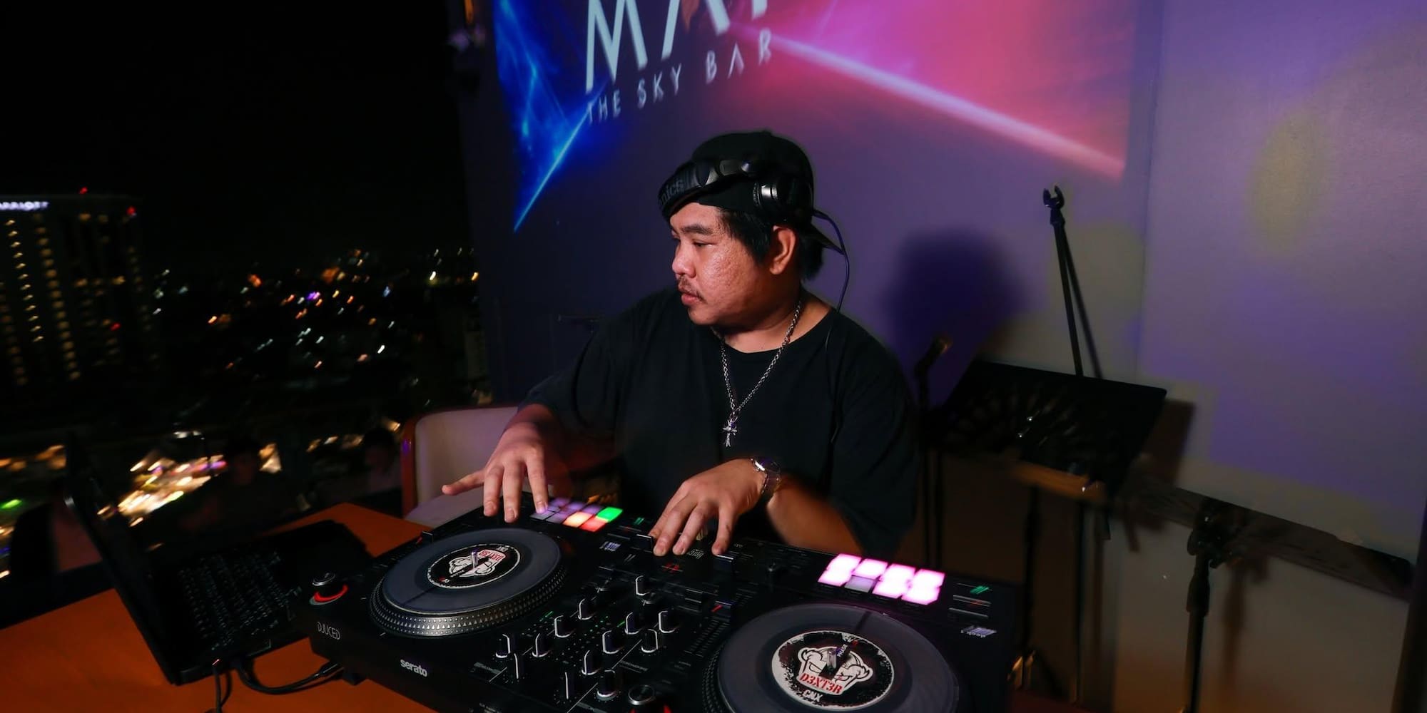 DJ with headphones mixing on controller at a rooftop sky bar at night.