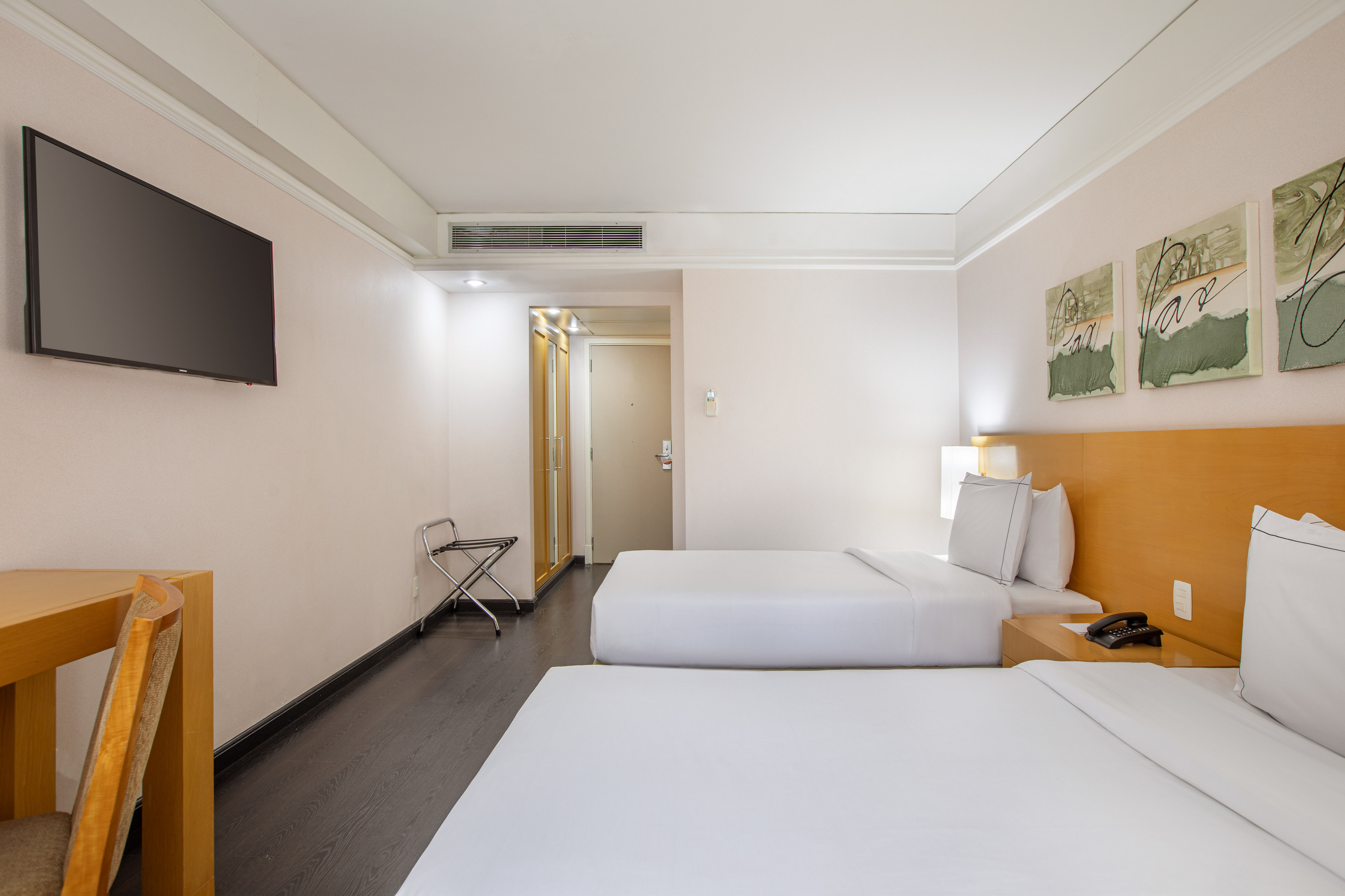 Bright hotel room with two beds, flat-screen TV, desk, and luggage rack.