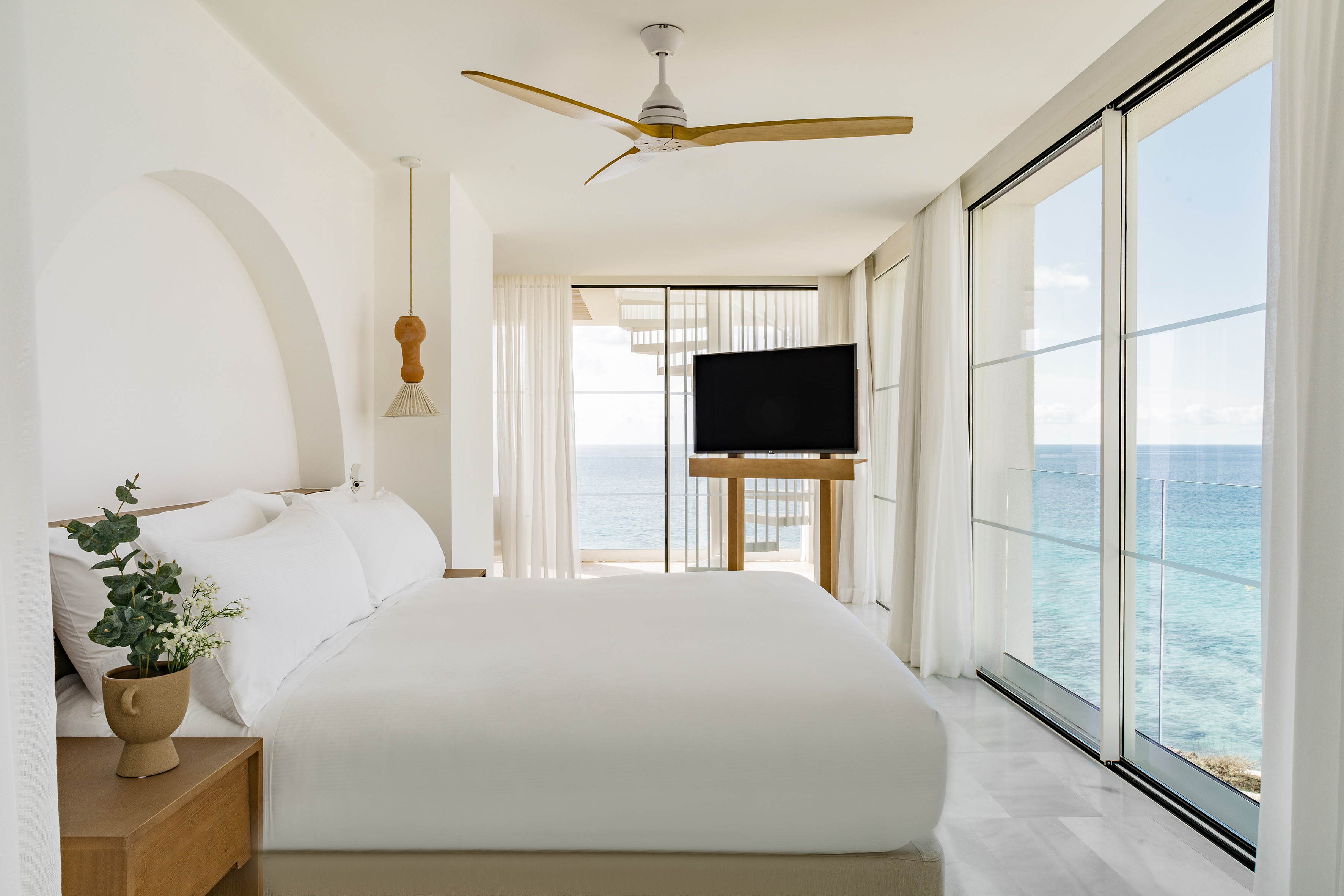 Coastal bedroom with white bed, ocean view, large glass doors, and wooden fan.