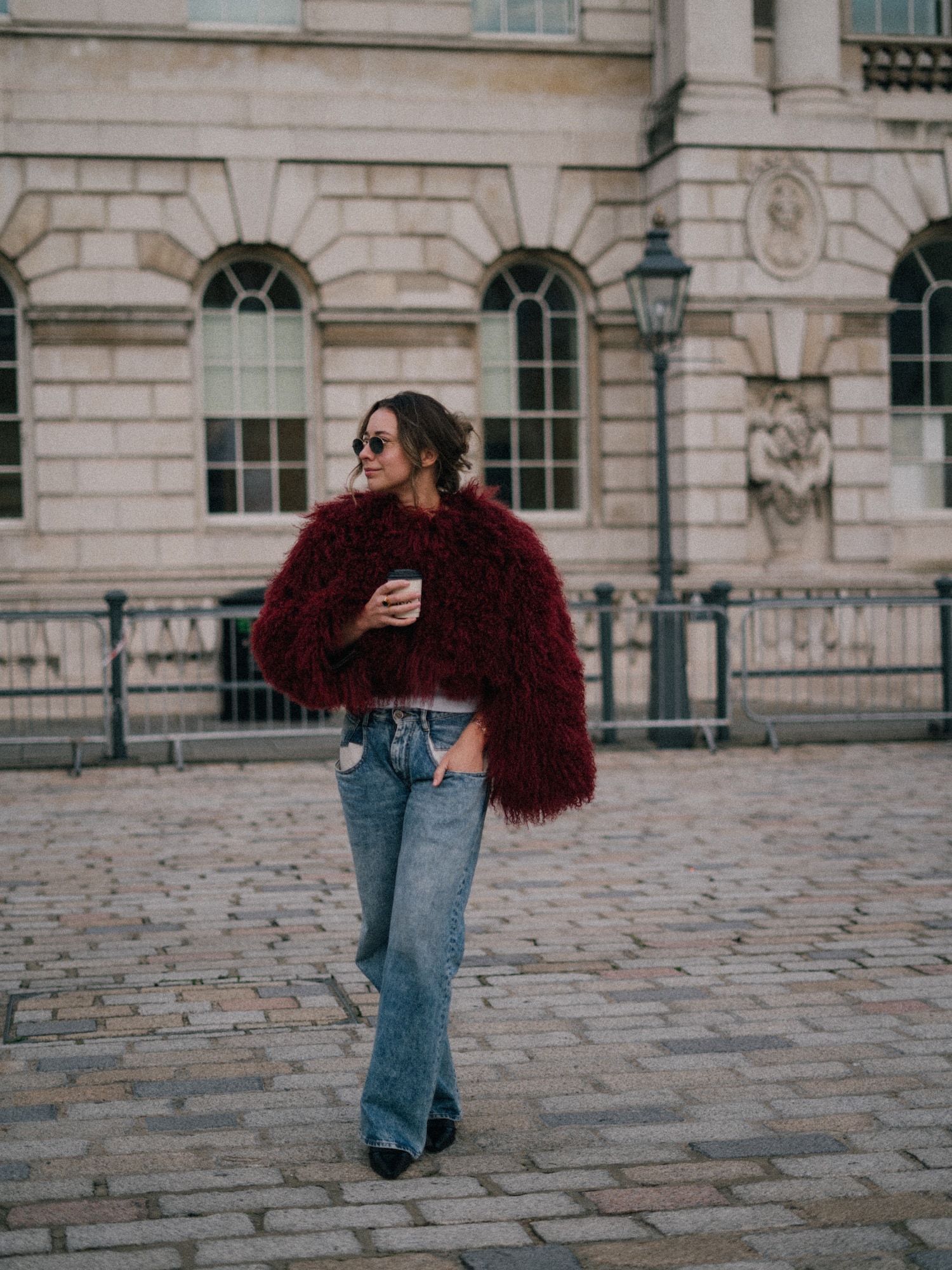 Woman in red faux fur coat, jeans, coffee, sunglasses, walking city street.