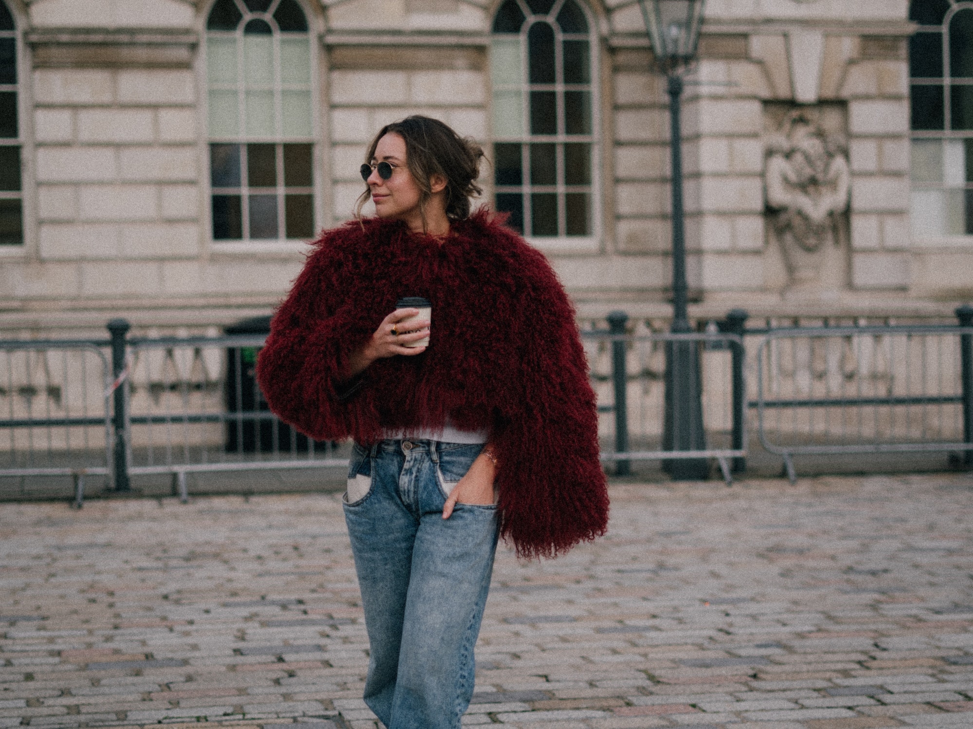 Woman in red faux fur coat, jeans, coffee, sunglasses, walking city street.