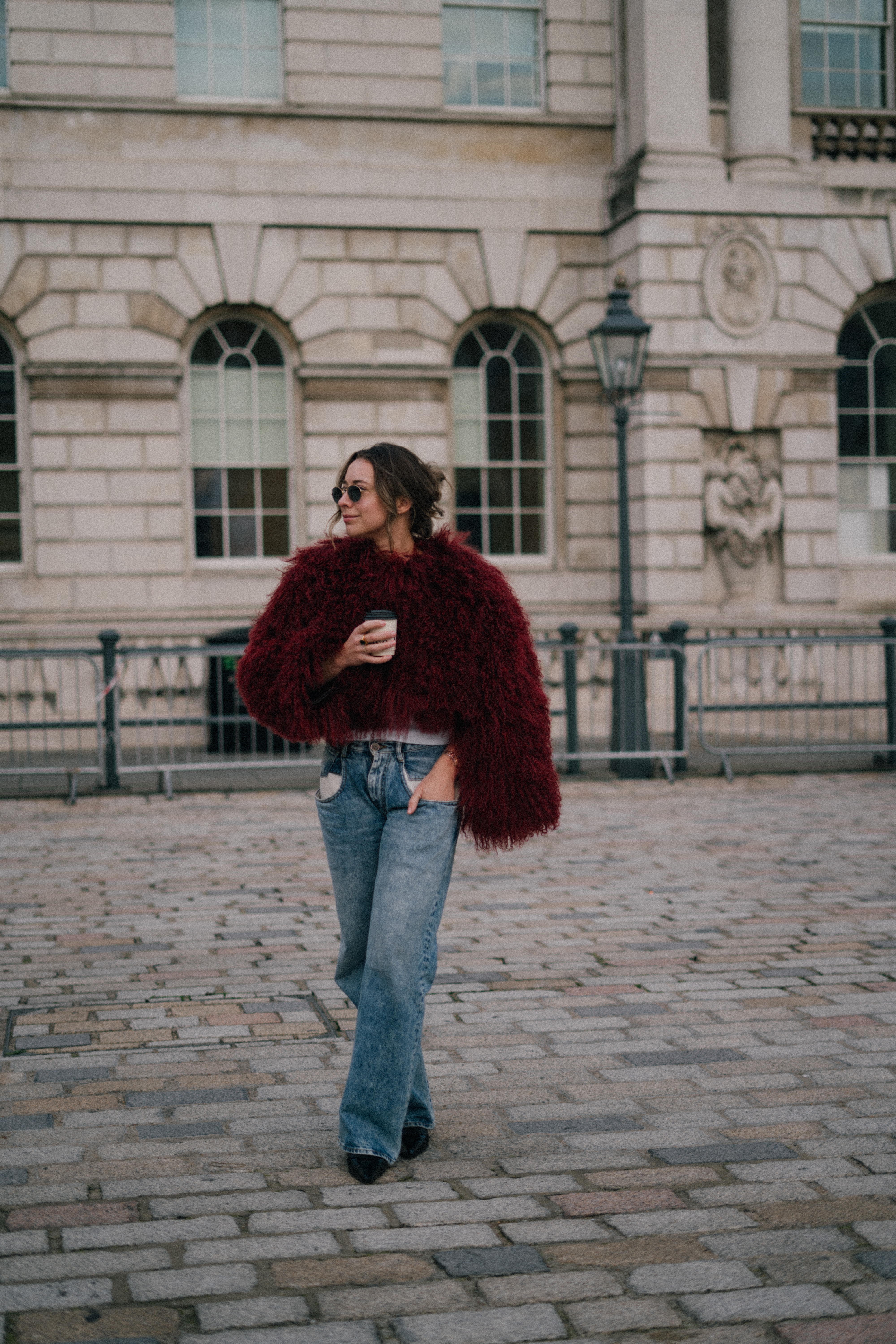 Woman in red faux fur coat, jeans, coffee, sunglasses, walking city street.