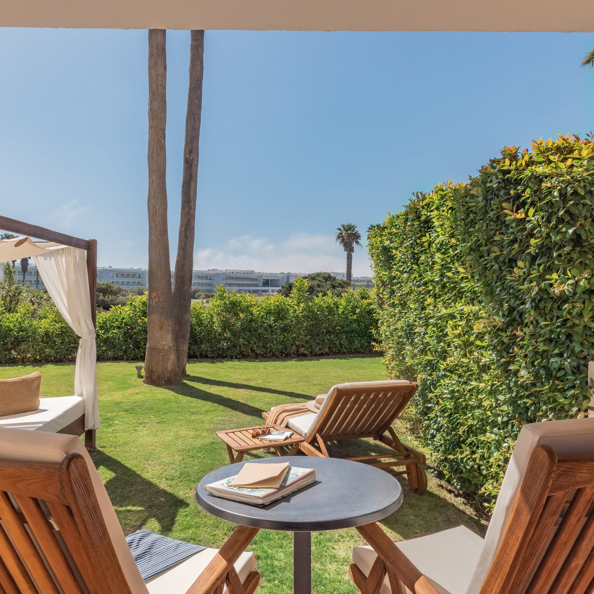 Outdoor hotel patio with garden, sunlounger, daybed, and palm trees.