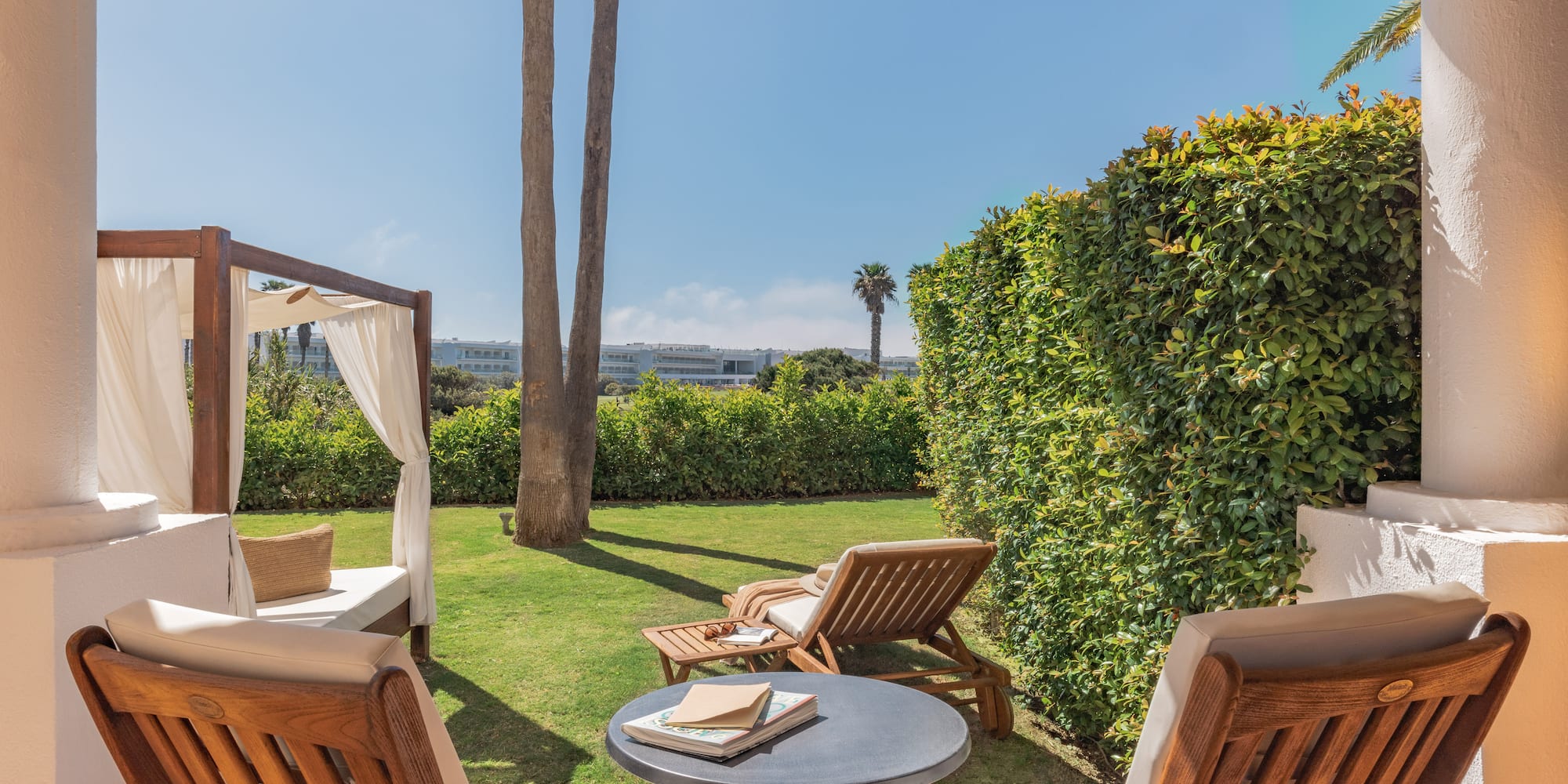 Outdoor hotel patio with garden, sunlounger, daybed, and palm trees.