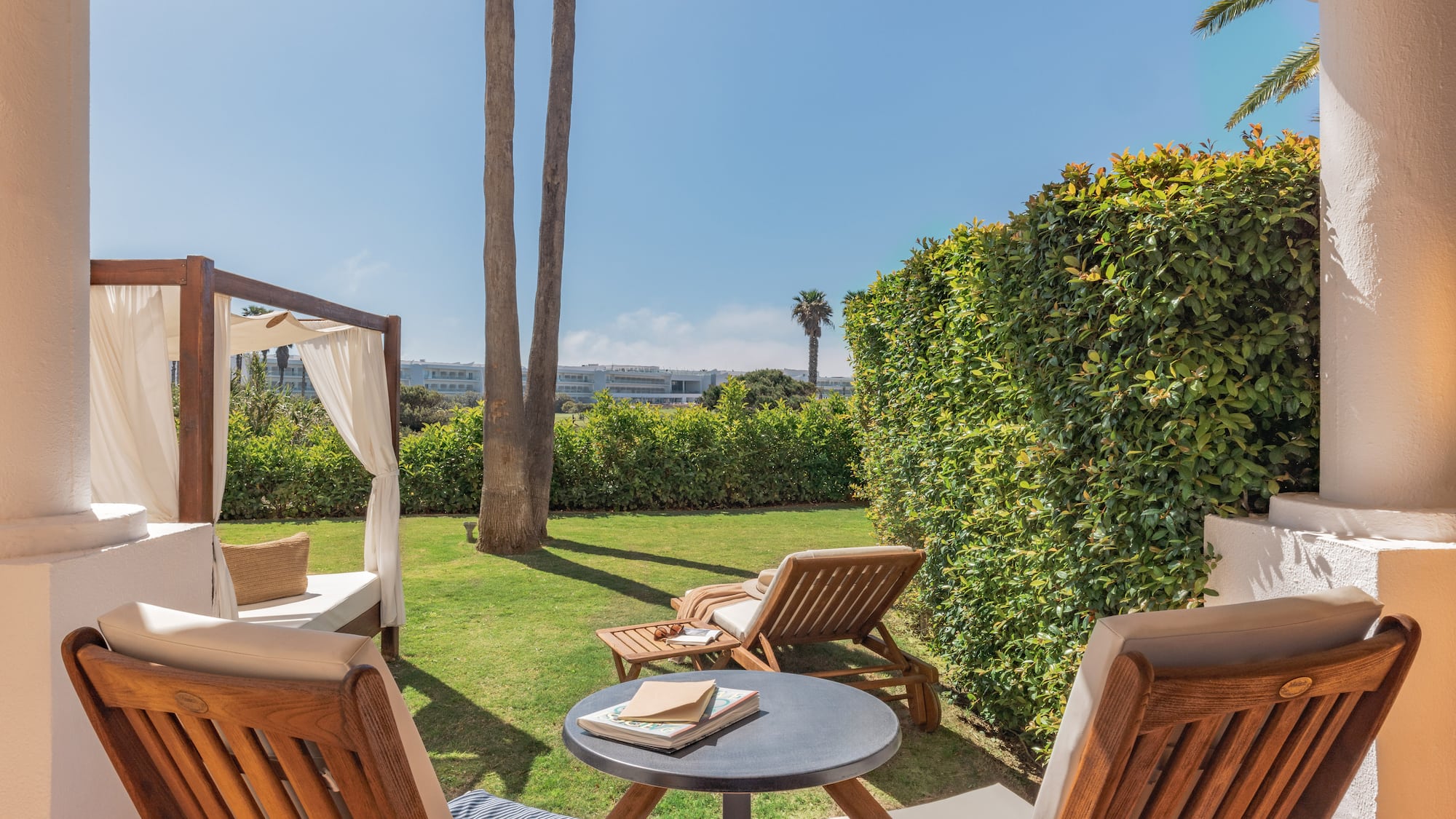Outdoor hotel patio with garden, sunlounger, daybed, and palm trees.