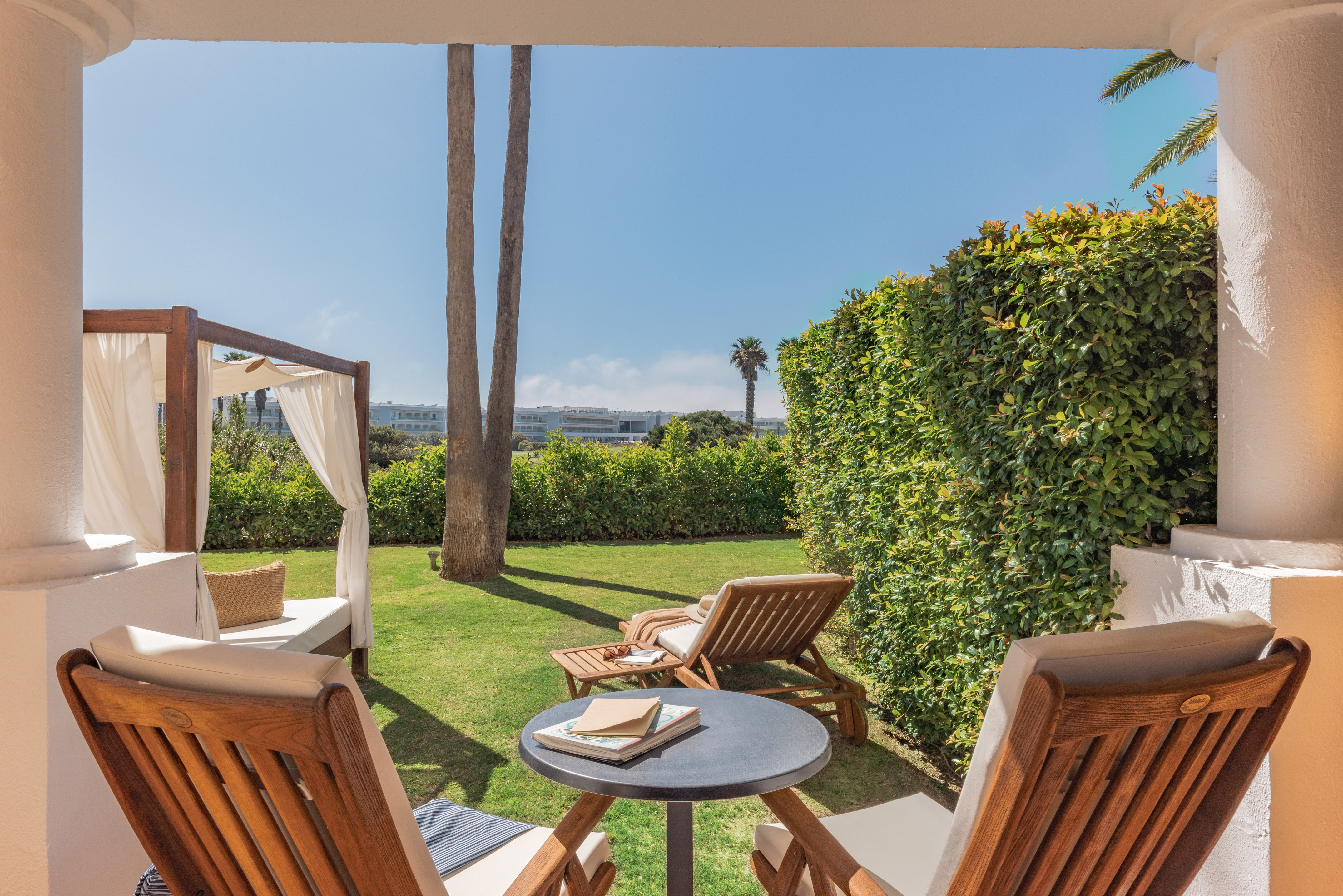Outdoor hotel patio with garden, sunlounger, daybed, and palm trees.