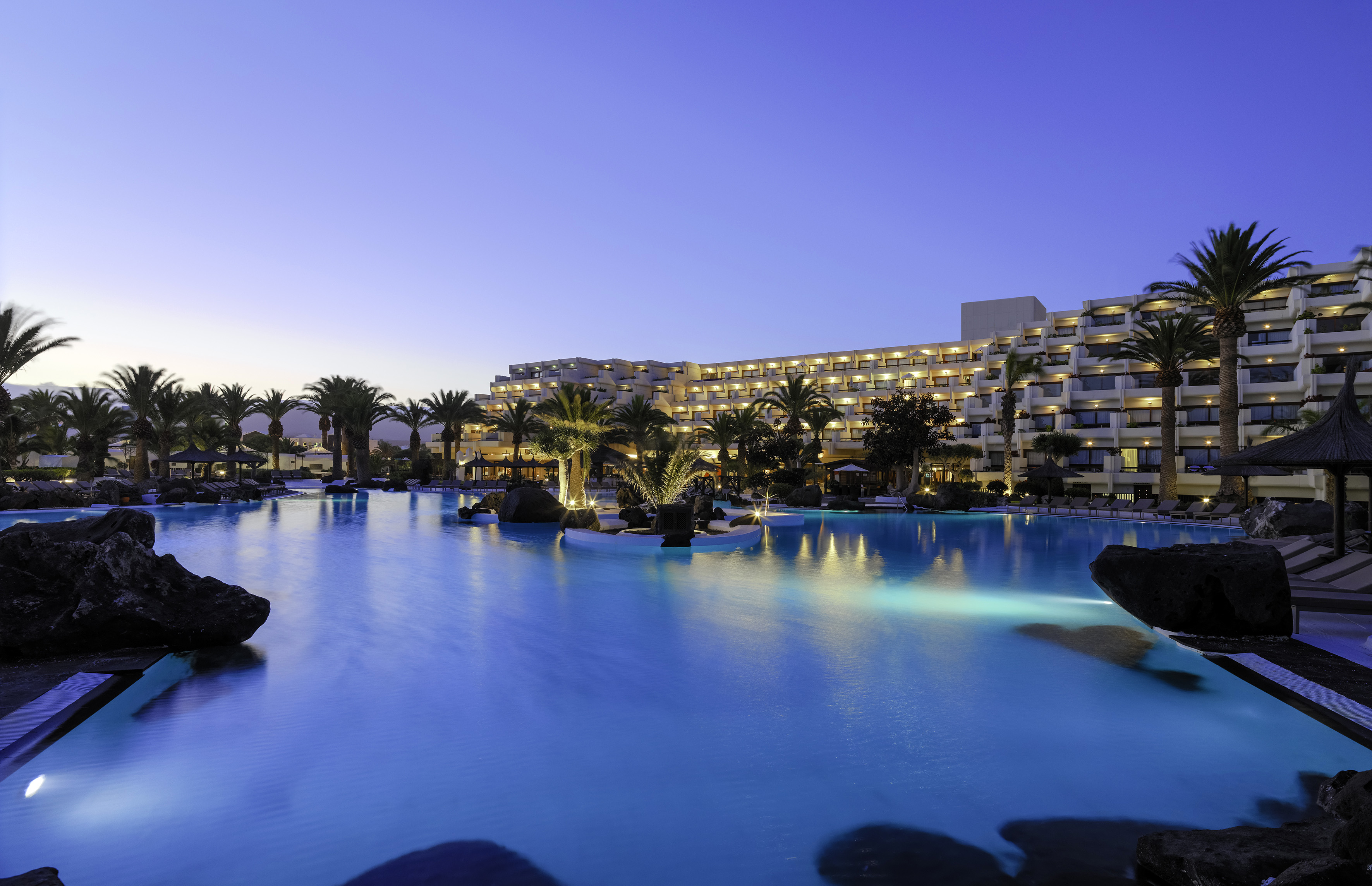 Luxury hotel with illuminated swimming pool and palm trees at dusk.