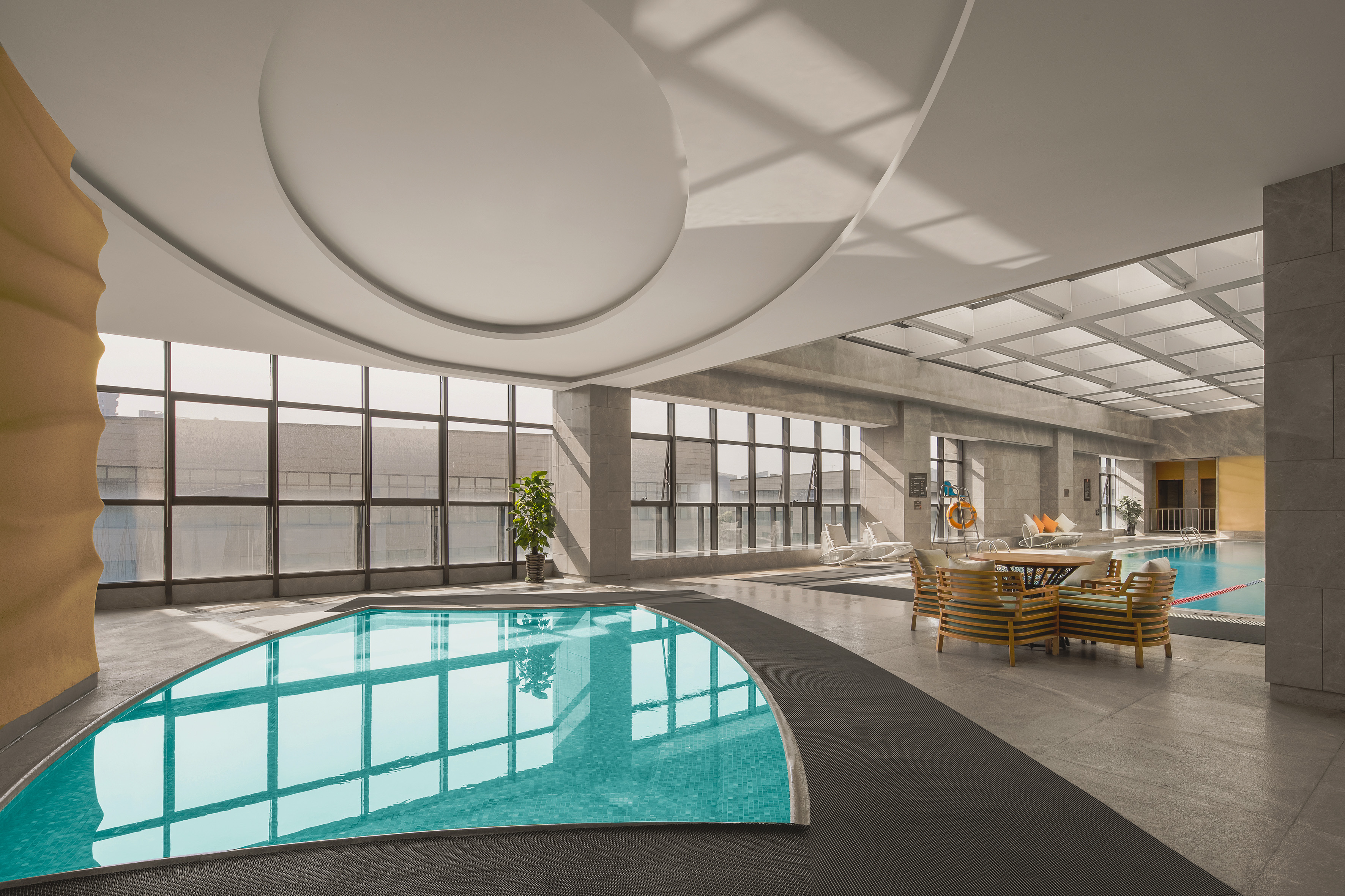 Modern indoor swimming pool with large windows and lounge area.