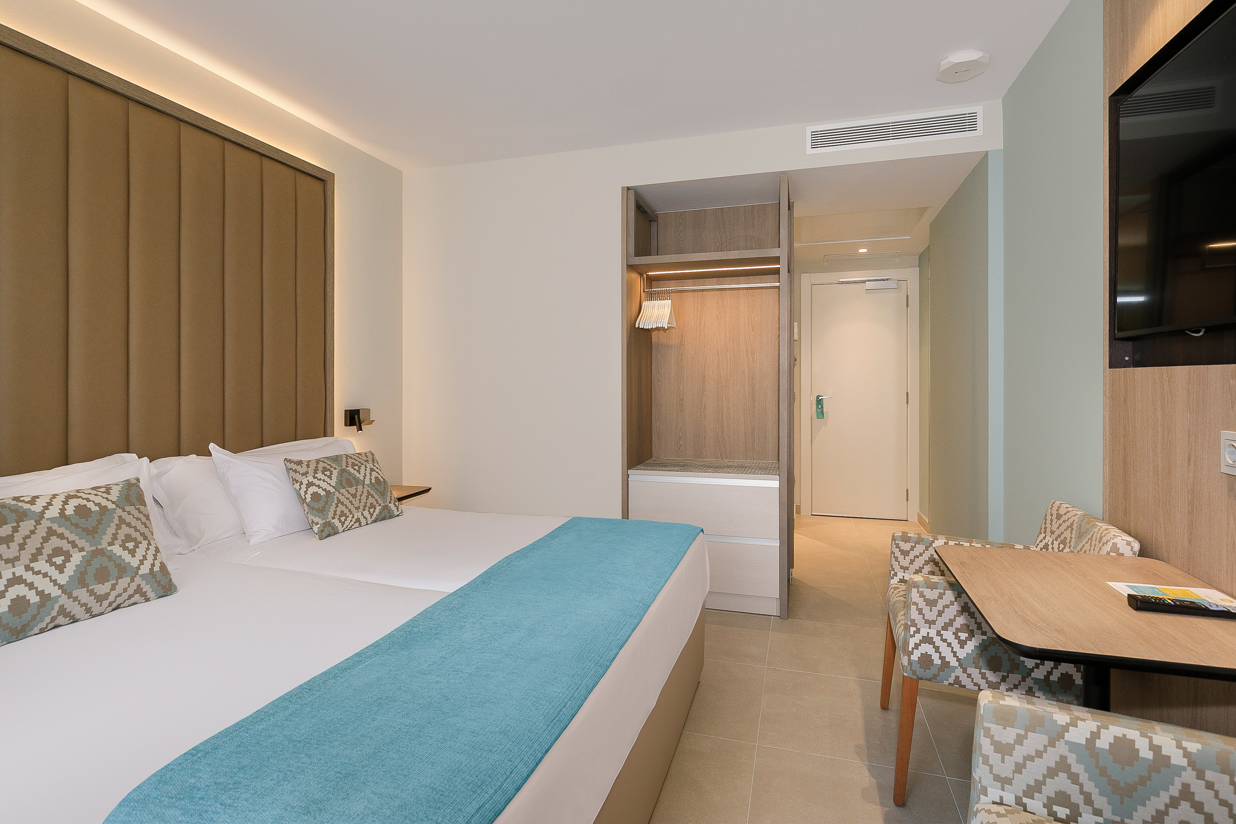 Modern hotel room with twin beds, open wardrobe, and dining table.