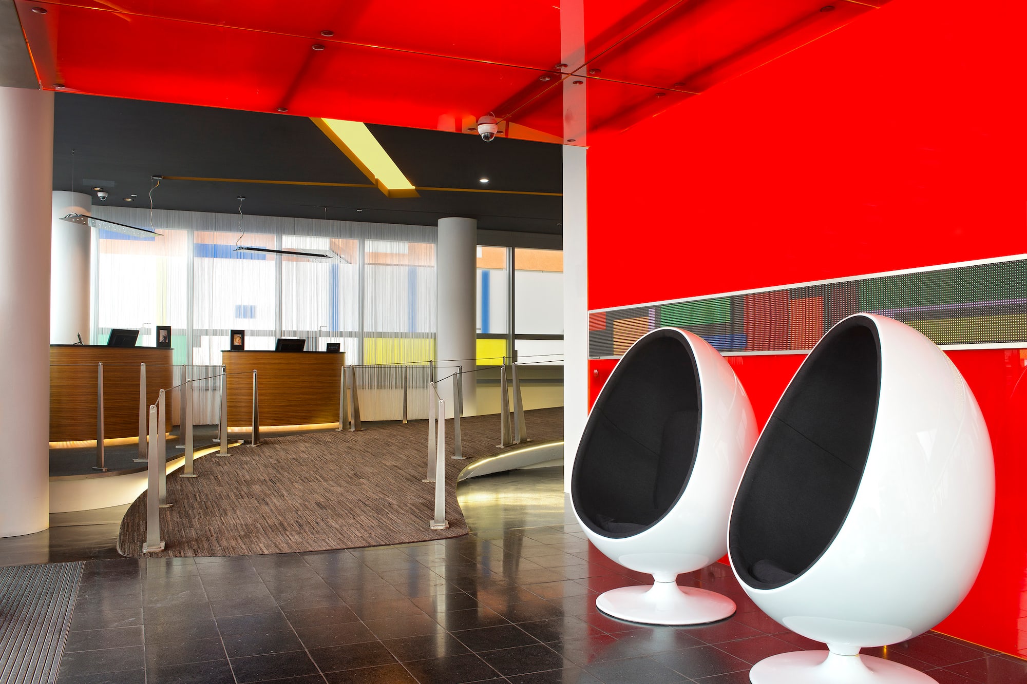 Modern hotel lobby with red walls, reception desks, and two white egg chairs.