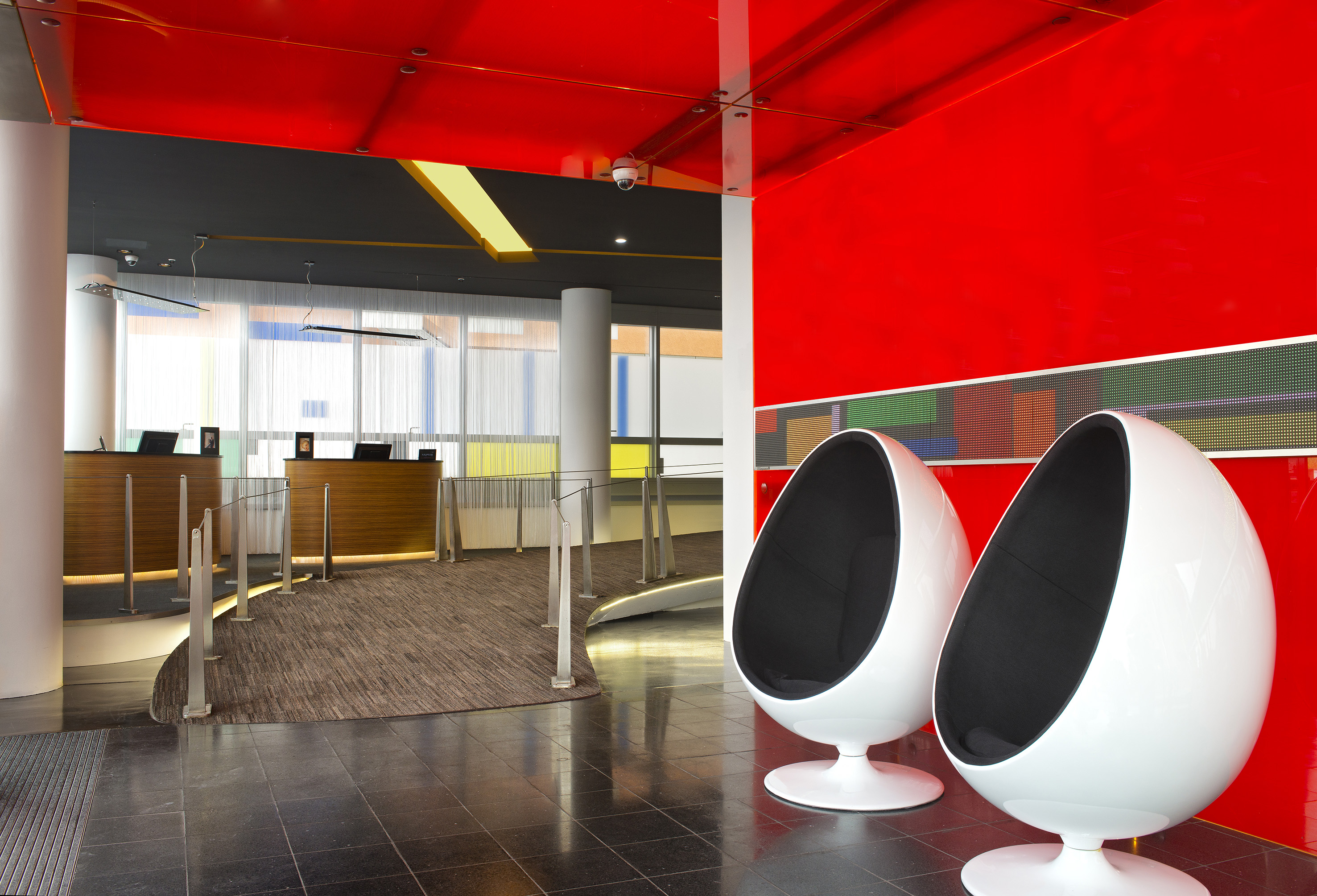 Modern hotel lobby with red walls, reception desks, and two white egg chairs.