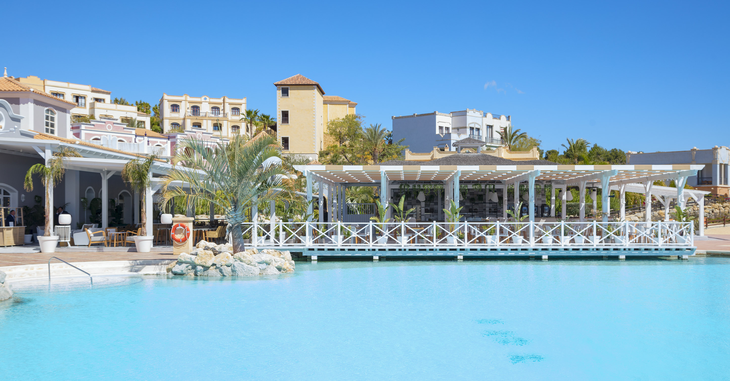 Sunny resort pool, outdoor dining pergola, and Mediterranean style hotel buildings.