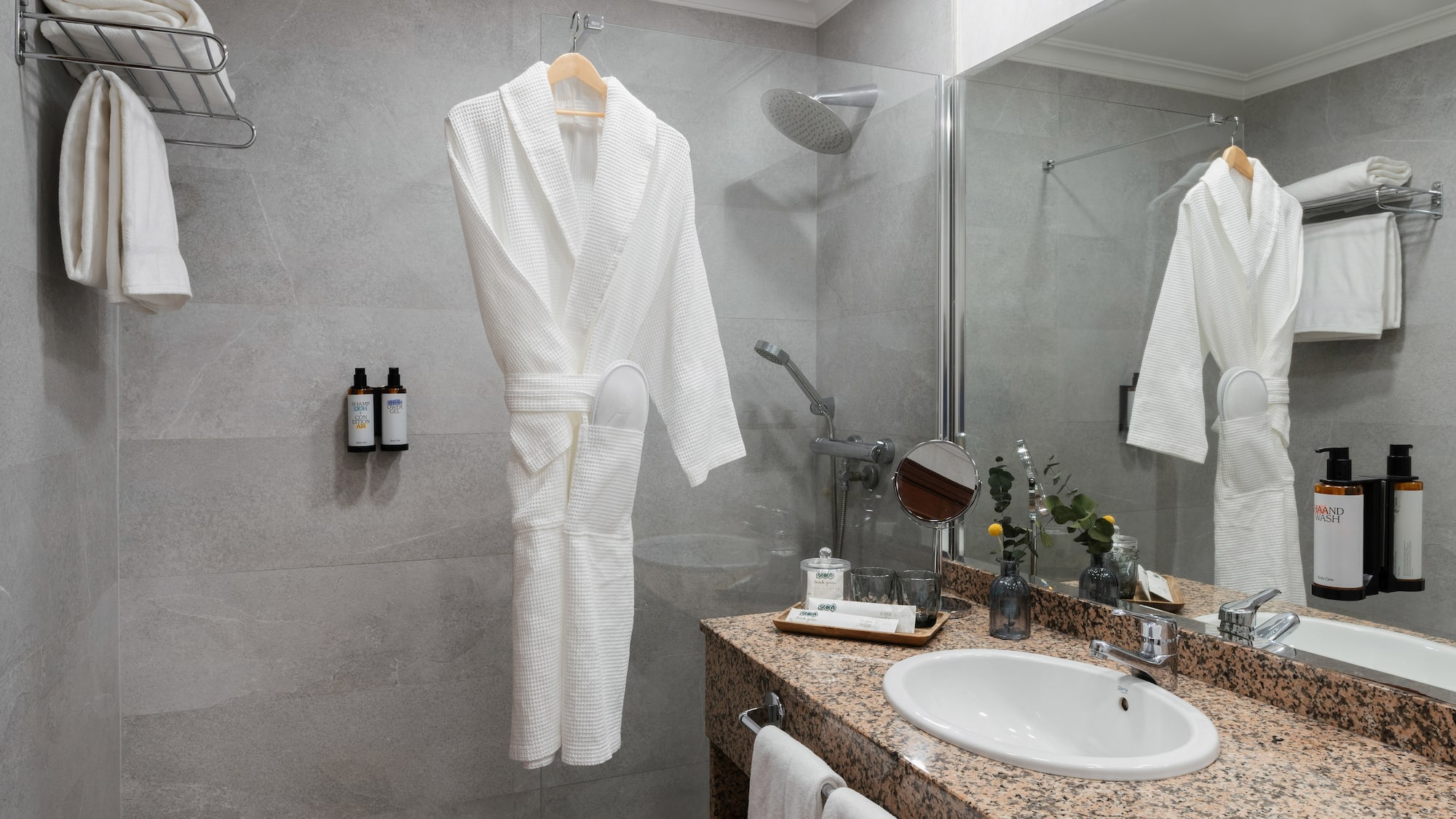 Modern hotel bathroom with shower, white bathrobe, granite vanity, and fresh towels.