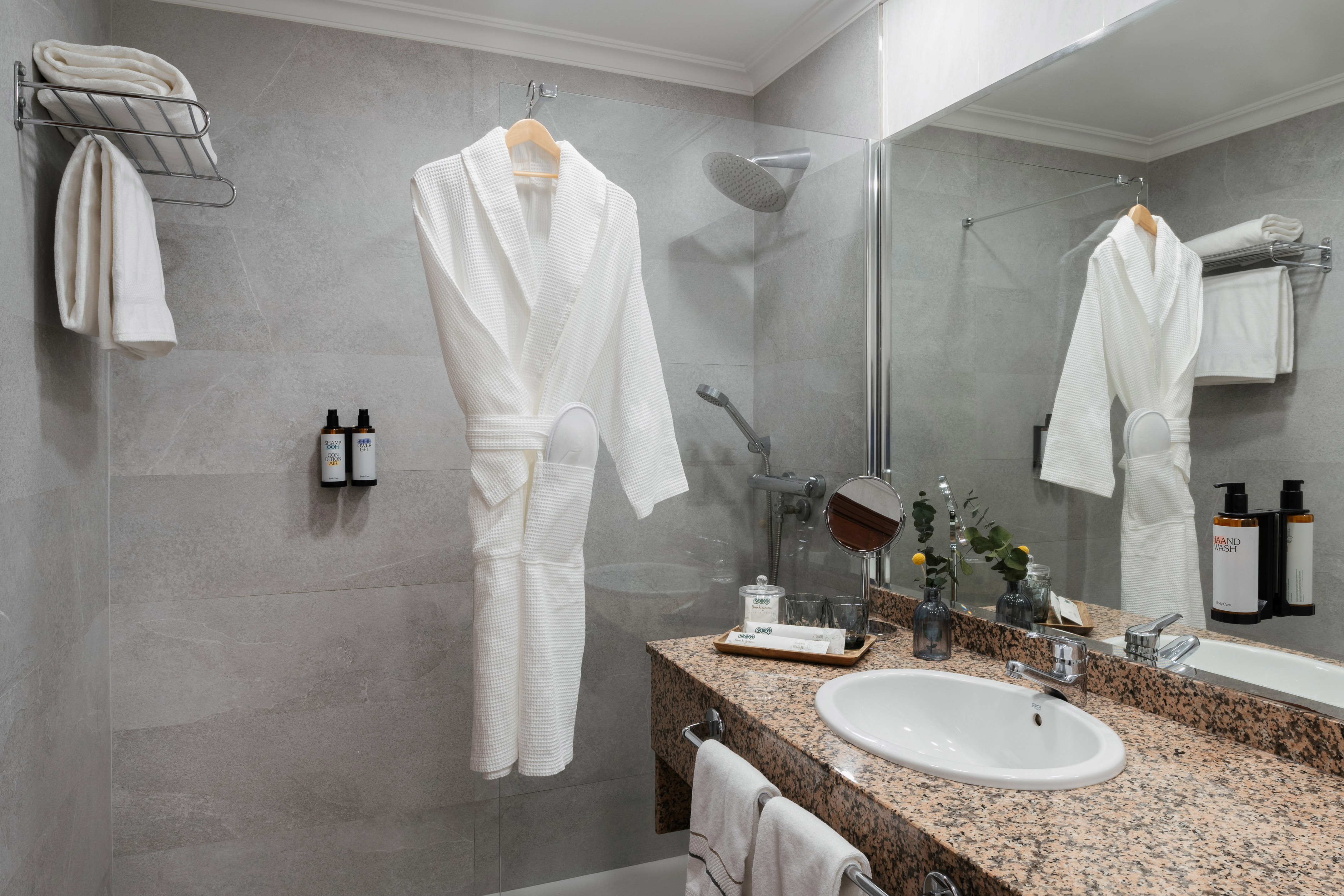 Modern hotel bathroom with shower, white bathrobe, granite vanity, and fresh towels.