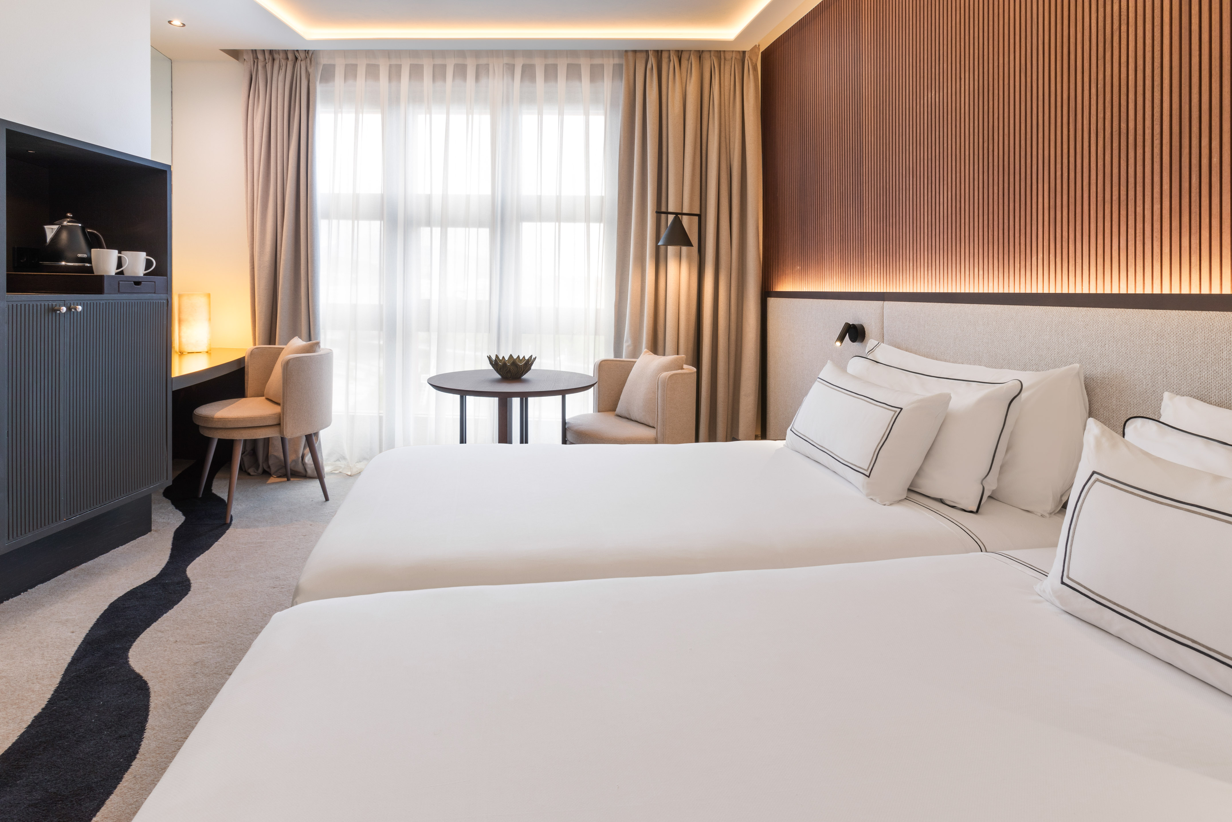 Modern hotel room with two twin beds, desk, window, and wood paneling.
