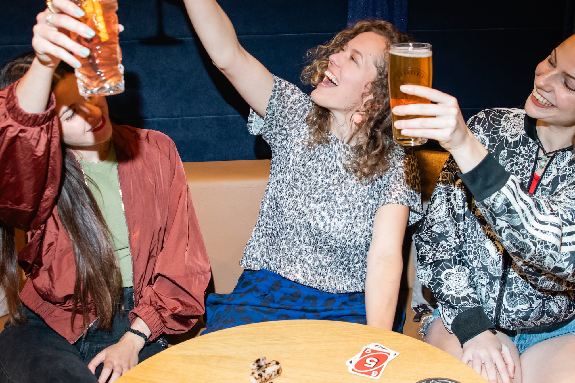 Tres amigas brindando con cócteles y cerveza en un bar, ambiente festivo.