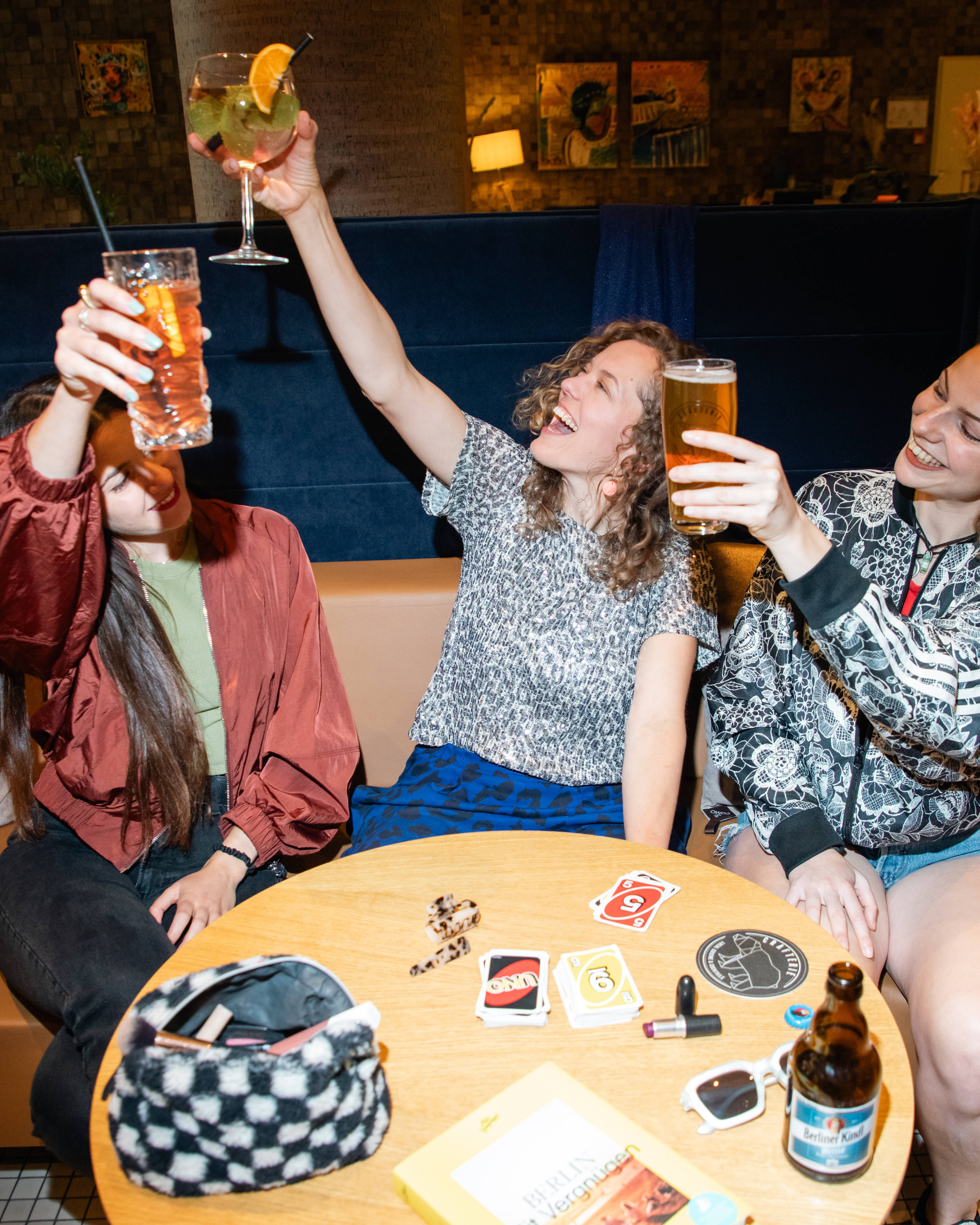 Tres amigas brindando con cócteles y cerveza en un bar, ambiente festivo.