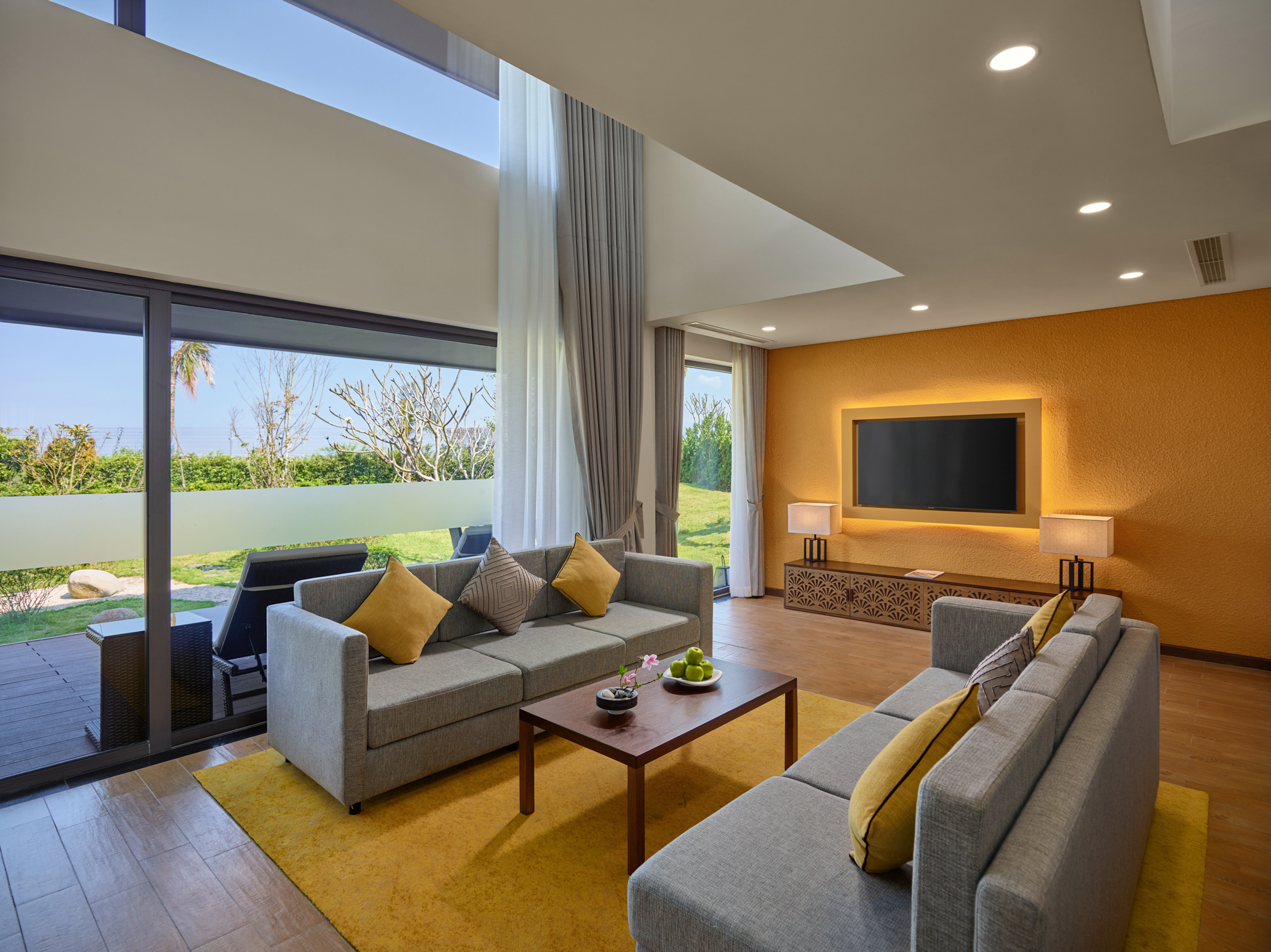 Modern living room with grey sofas, yellow rug, large windows, and patio view.