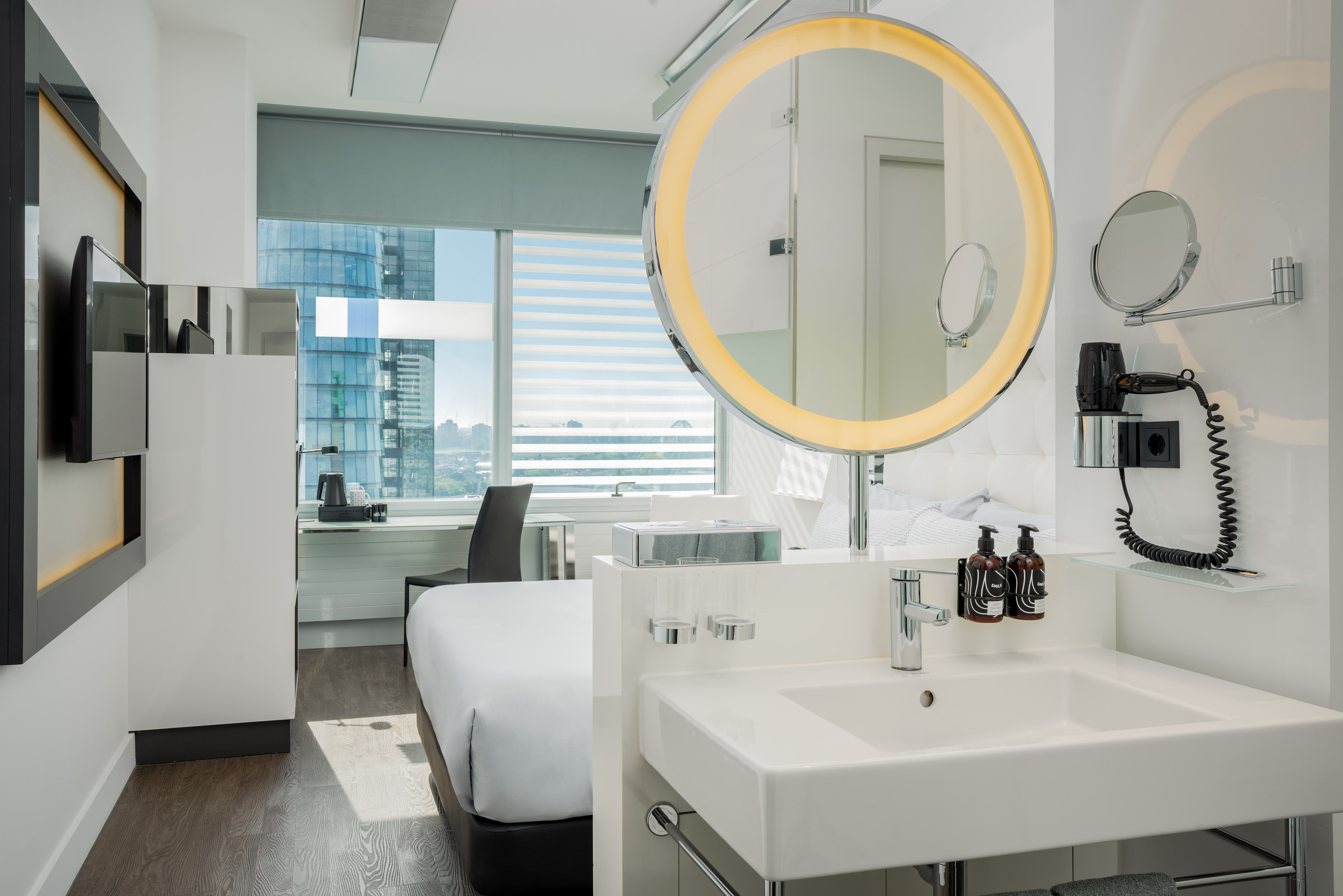 Sleek hotel room with illuminated mirror, vanity, bed, and city view.