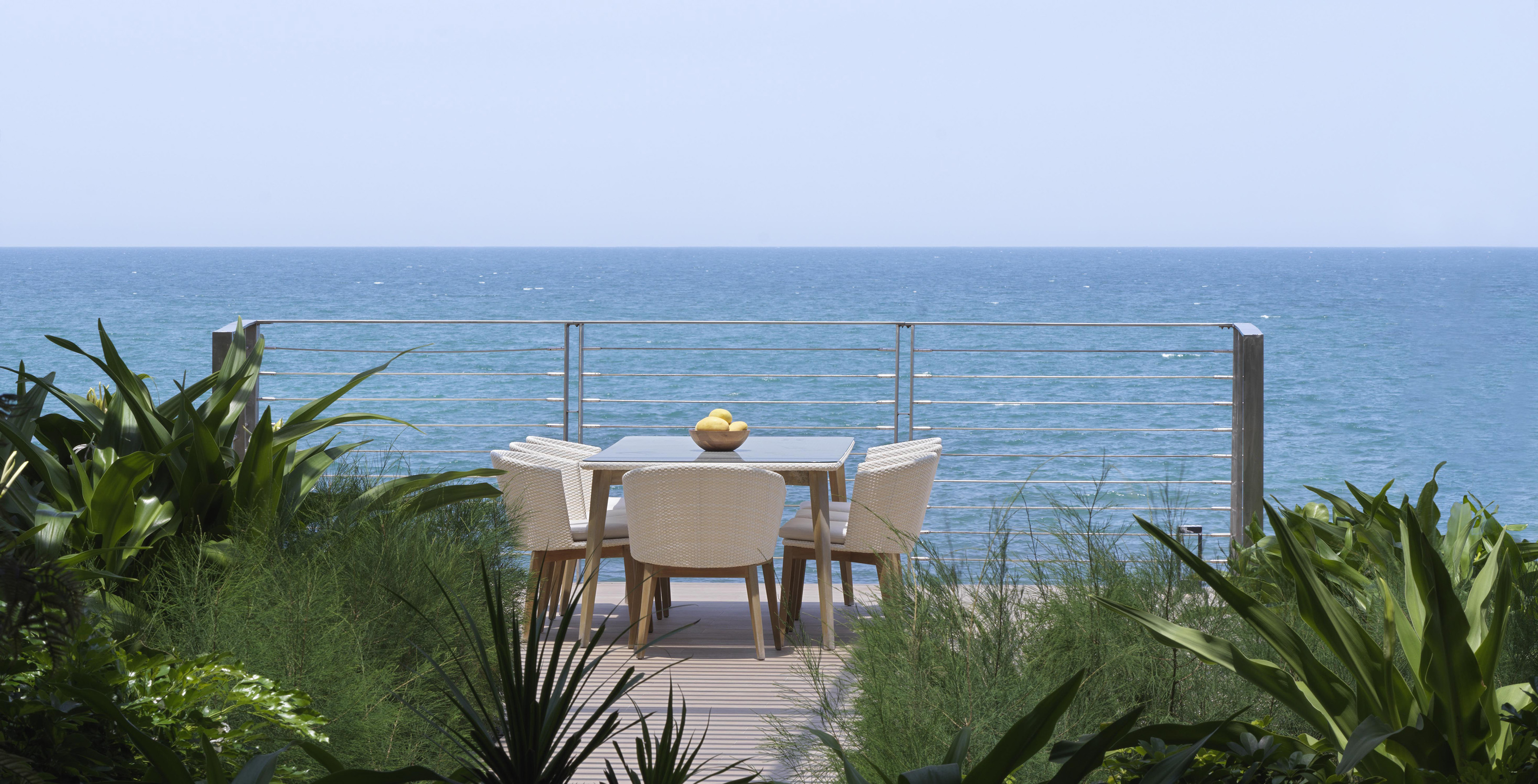Outdoor dining deck with ocean view, tropical plants, and rattan furniture.