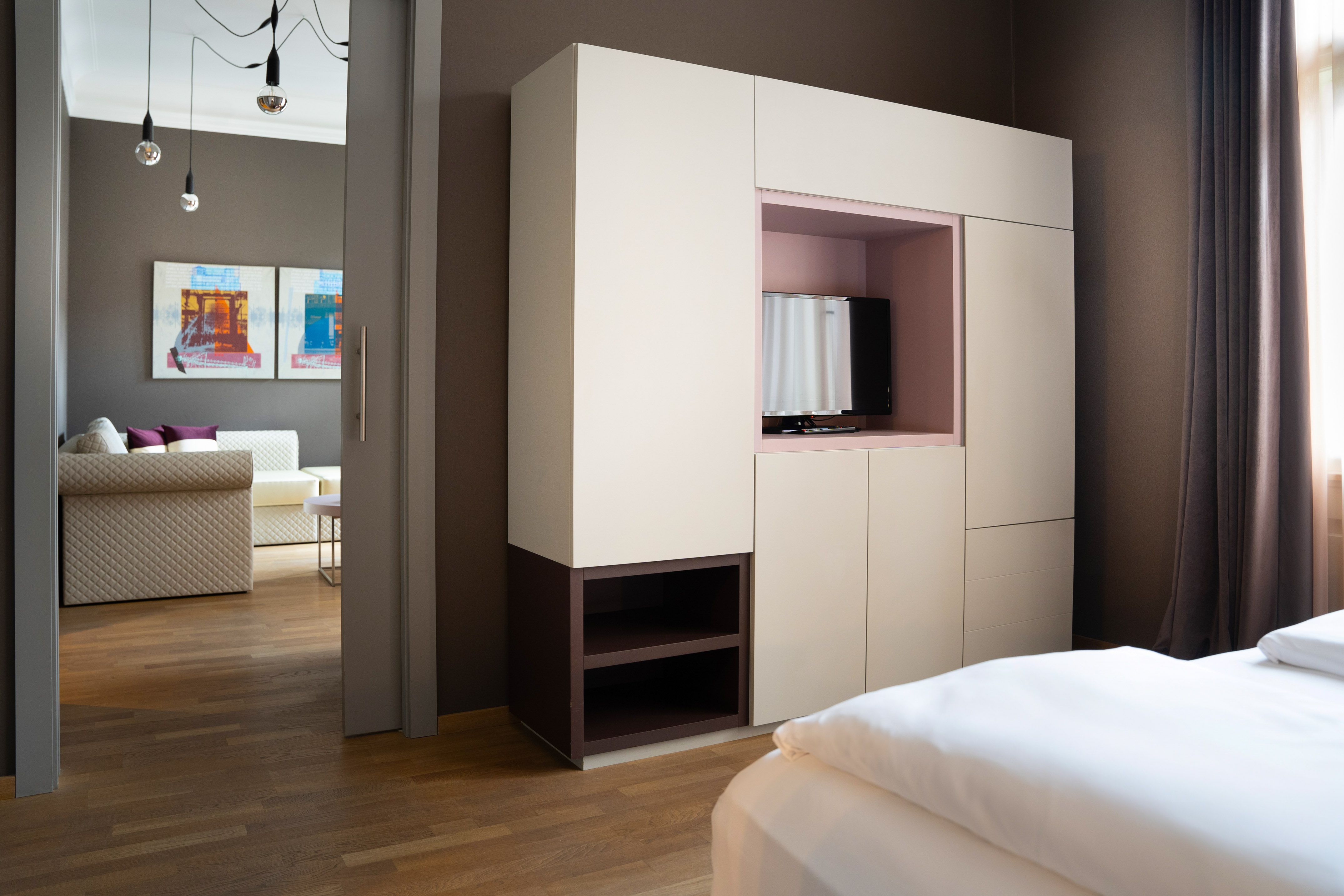 Modern bedroom with built-in TV wardrobe, white bed, wooden floor, and living area.