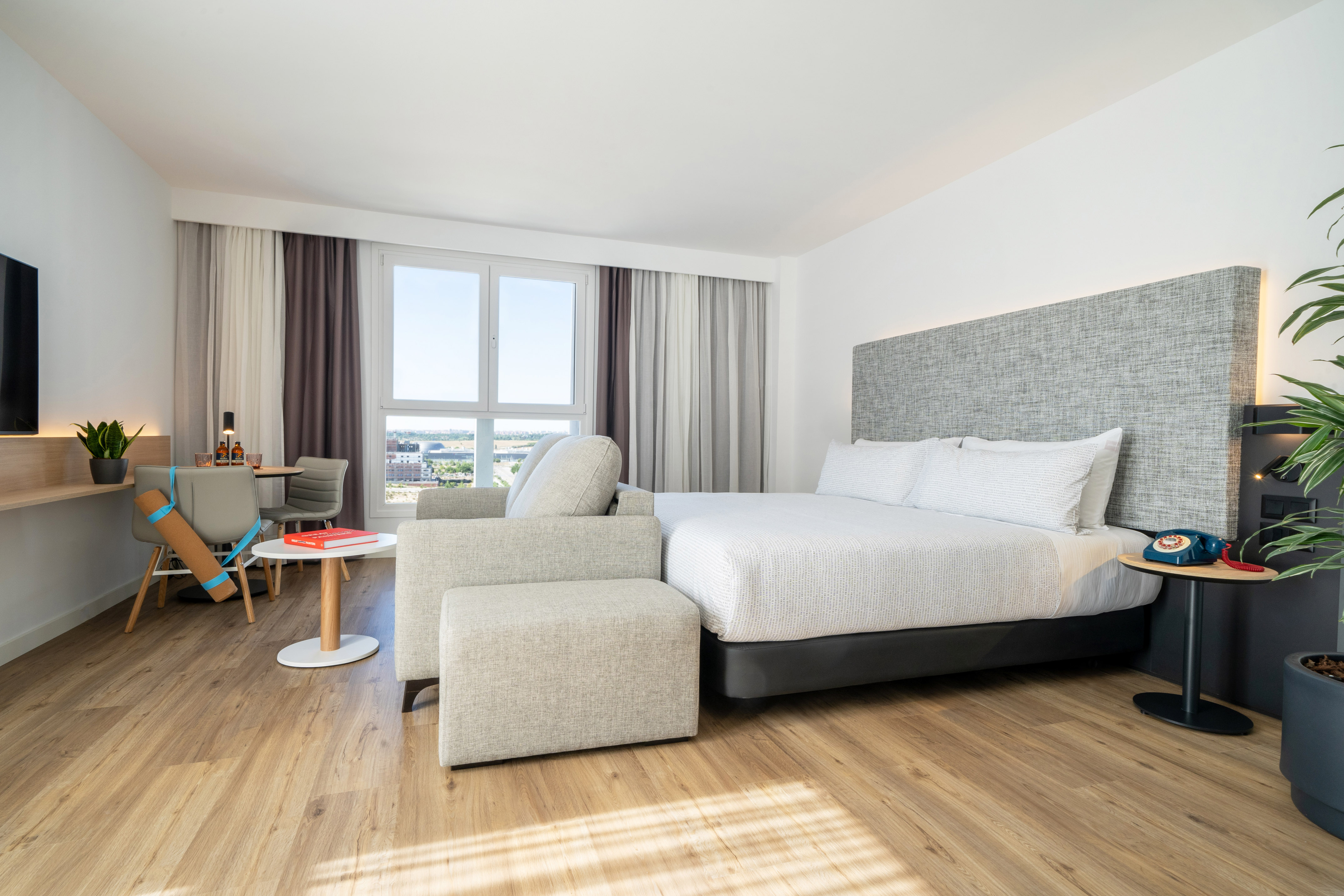 Bright modern hotel studio with bed, sofa, dining area, and wooden floor.