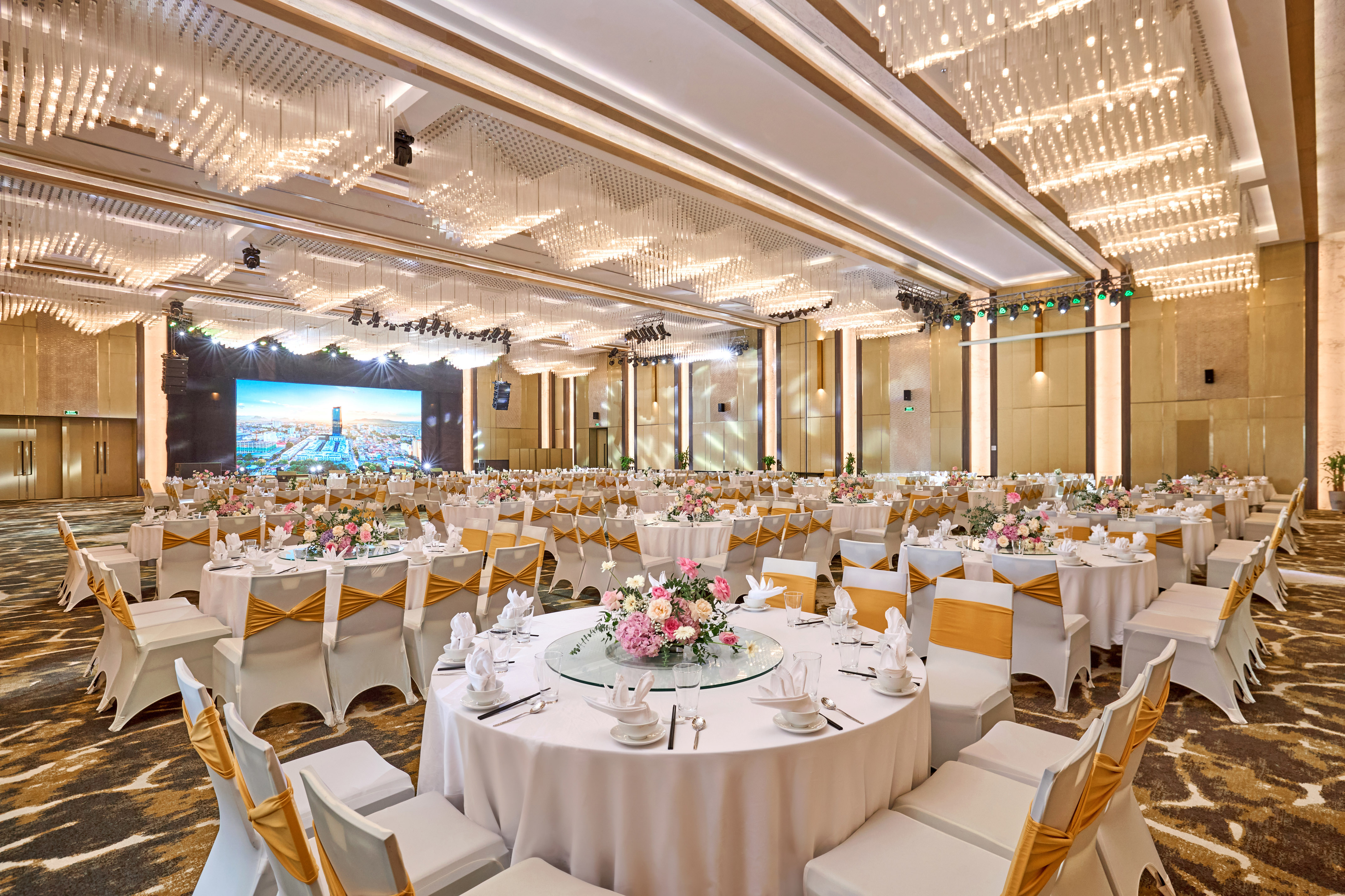 Elegant ballroom with set dining tables, gold chairs, floral centerpieces, and screen.