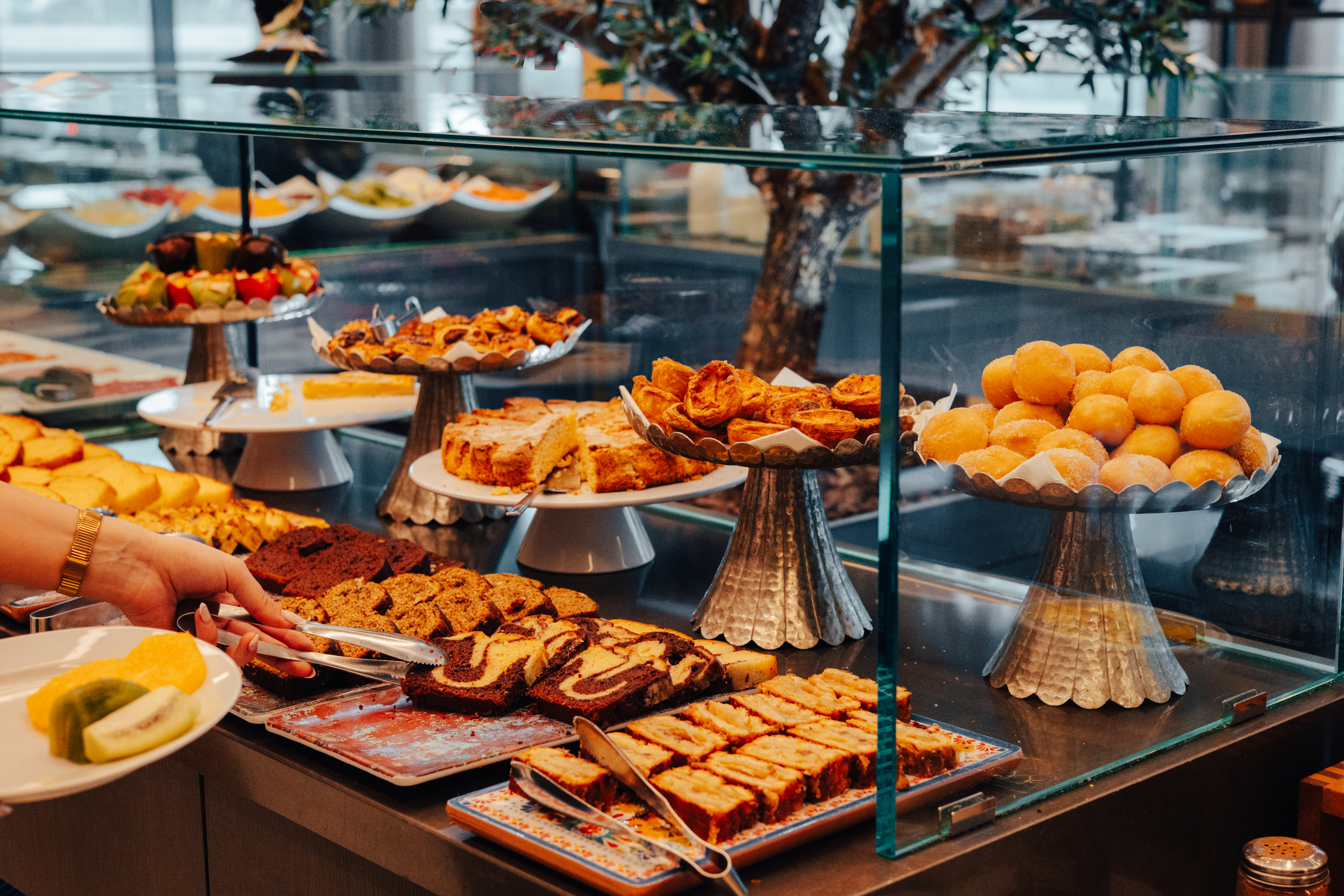 Hand choosing cake at hotel breakfast buffet with various pastries & desserts.