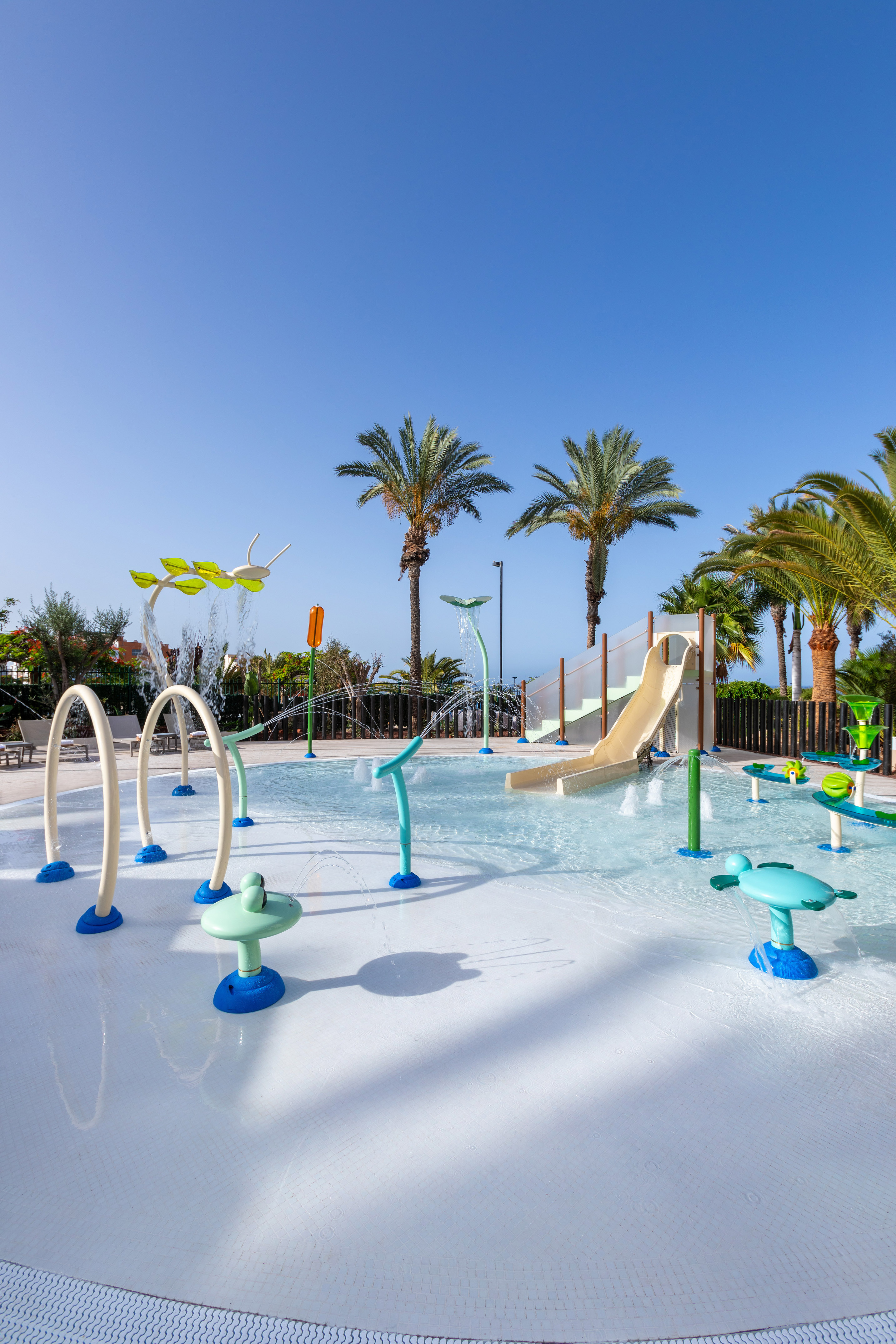 Outdoor kids' splash pad with water features, slide, and palm trees.
