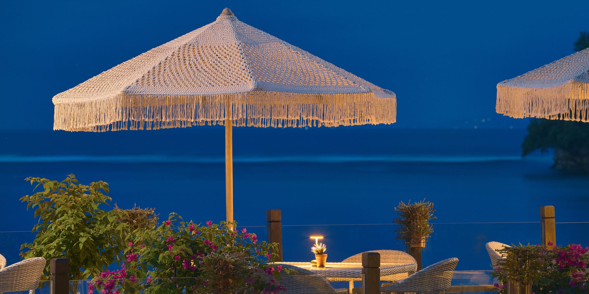 Outdoor dining terrace with macrame umbrellas, sea view at dusk.