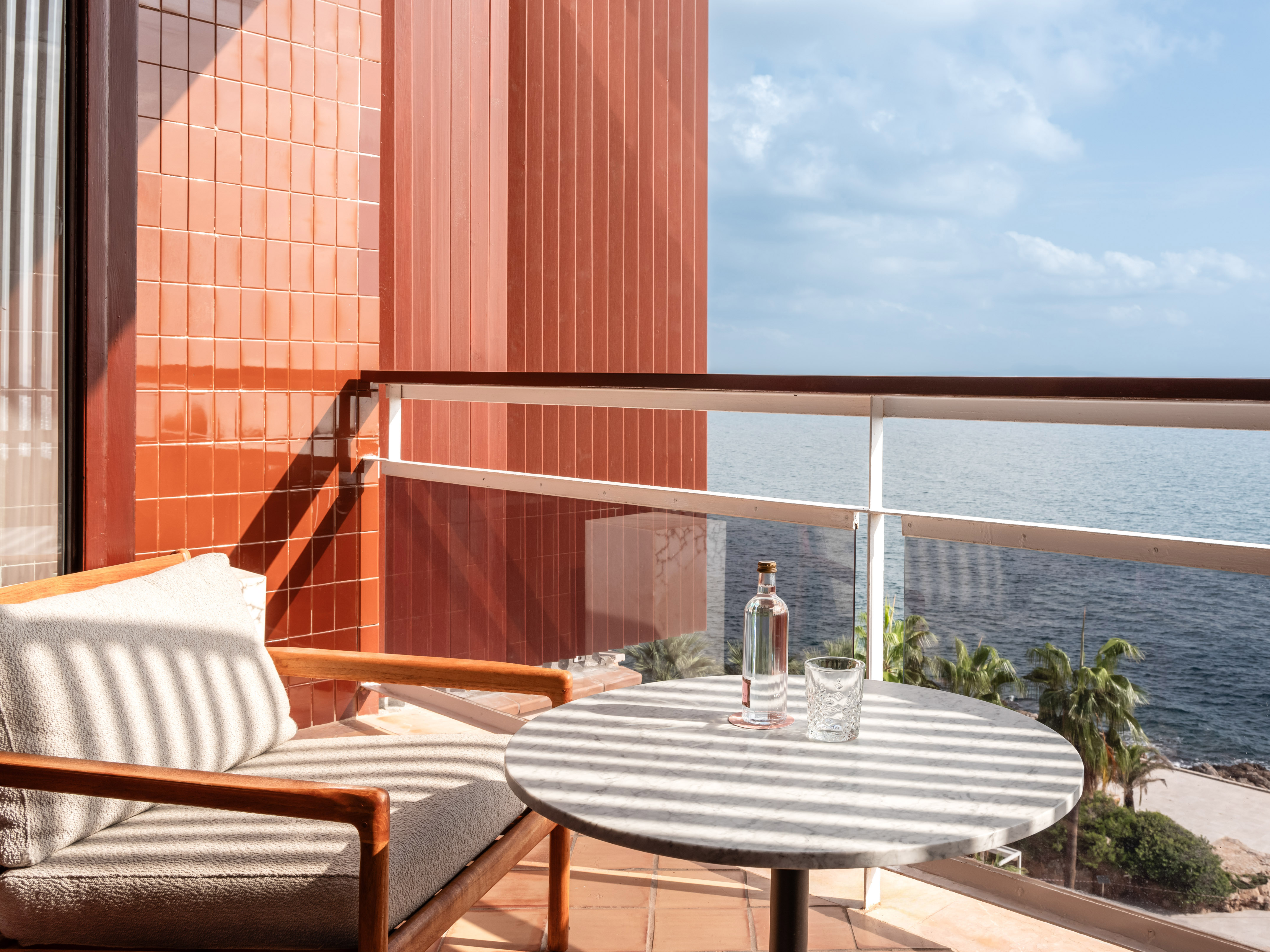 Hotel balcony with marble table, chair, and ocean view over palm trees.