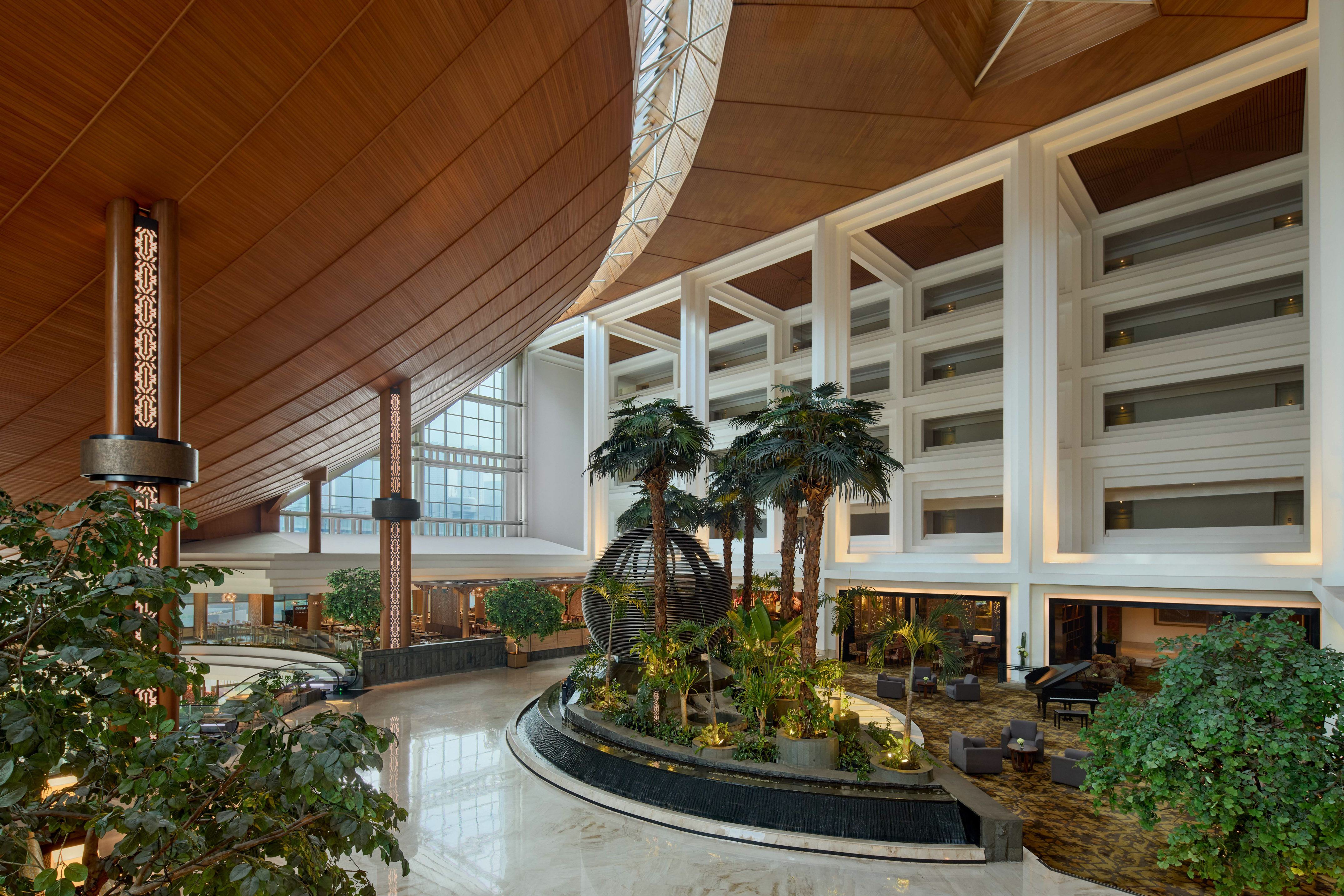 Luxurious hotel lobby atrium with curved wooden ceiling, palm trees, water feature.