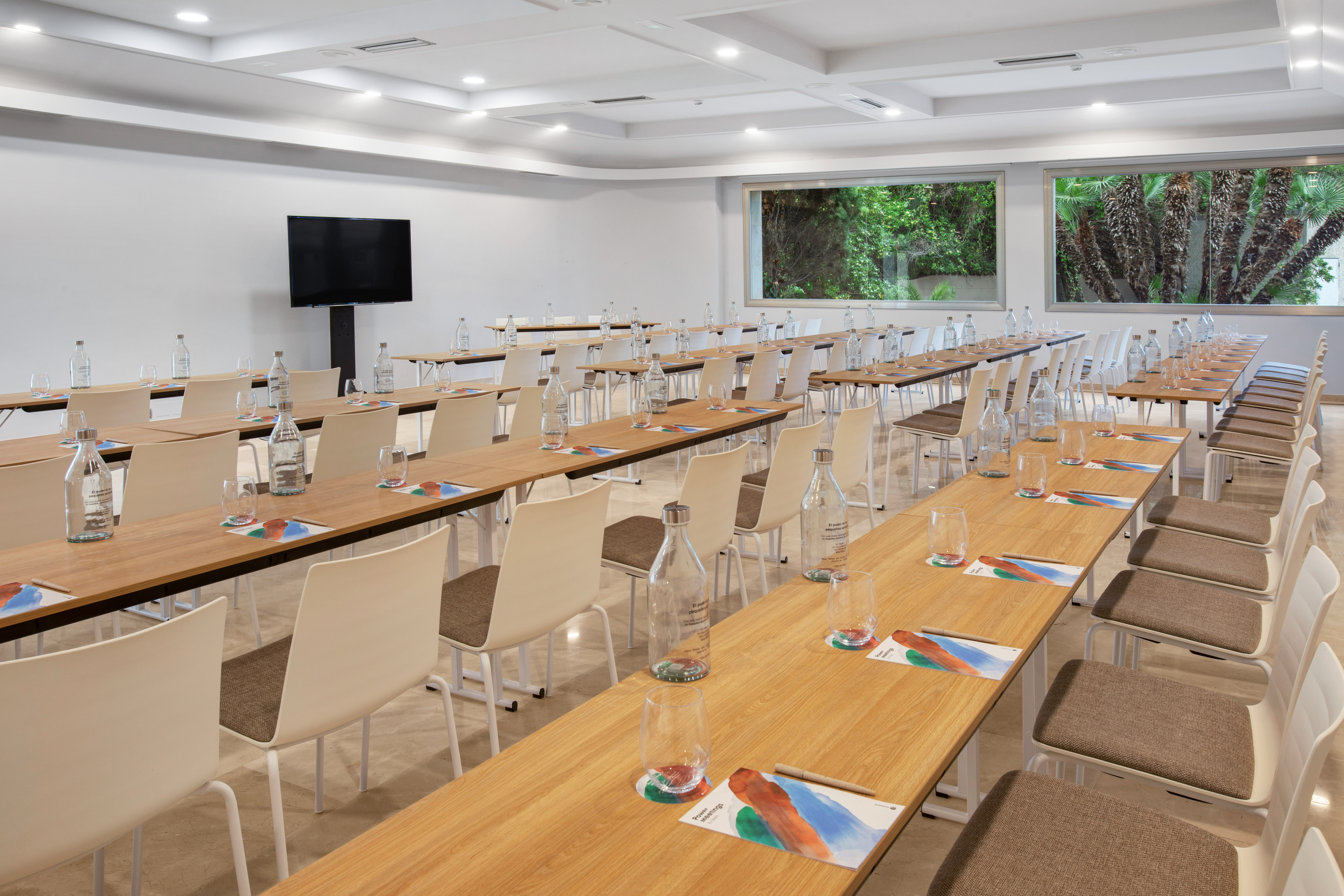 Conference room with wooden tables, white chairs, TV, and green views outside.