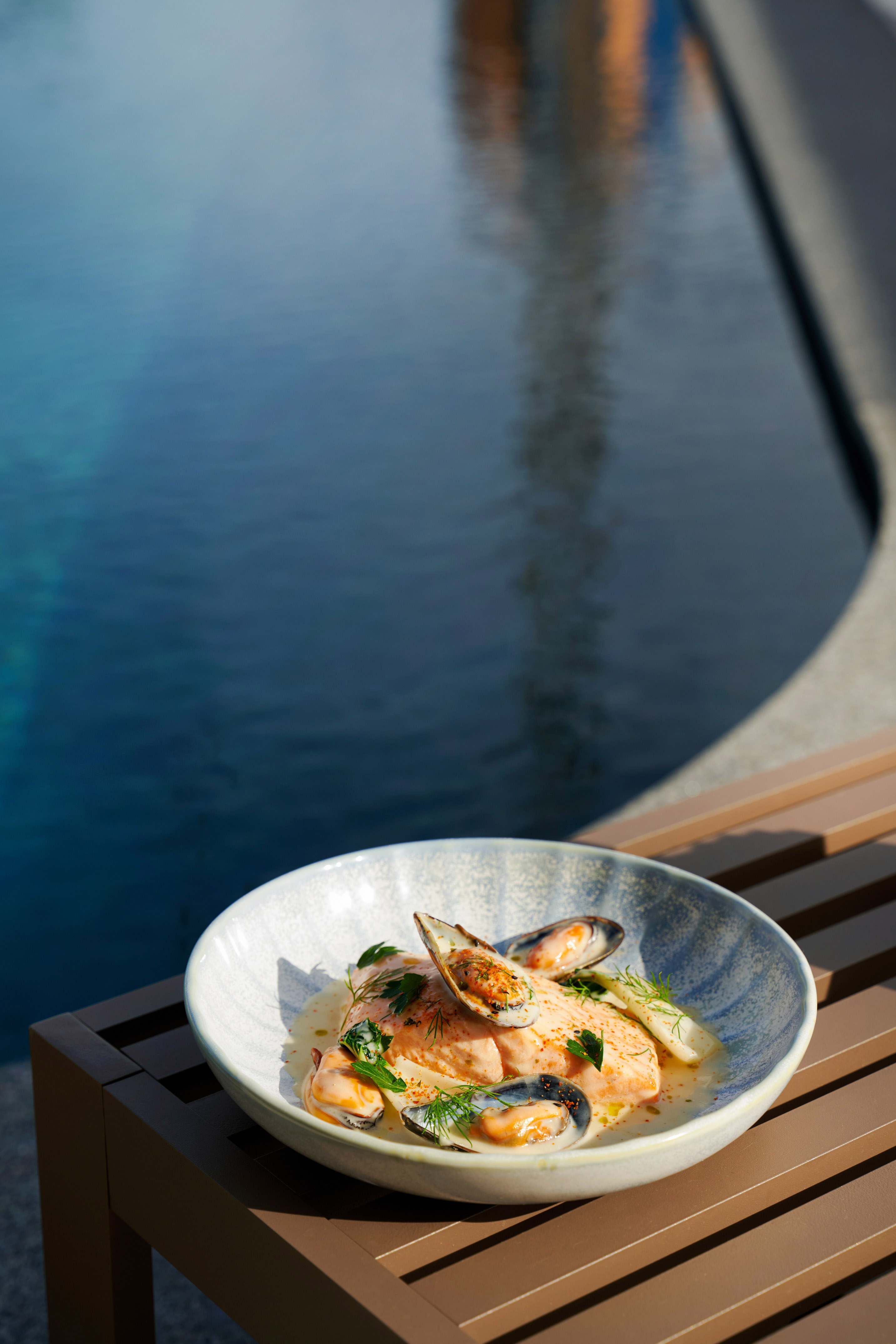 Salmon and mussels with herbs in a bowl, by a blue pool.