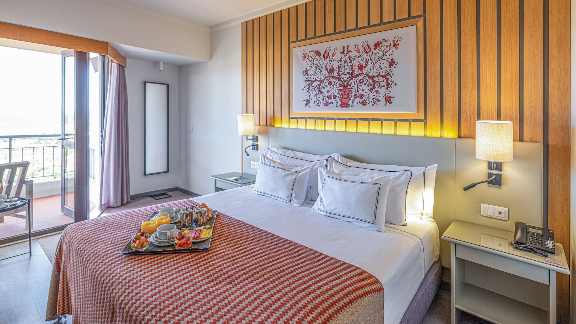 Hotel room with breakfast tray on bed, balcony, and wood wall decor.