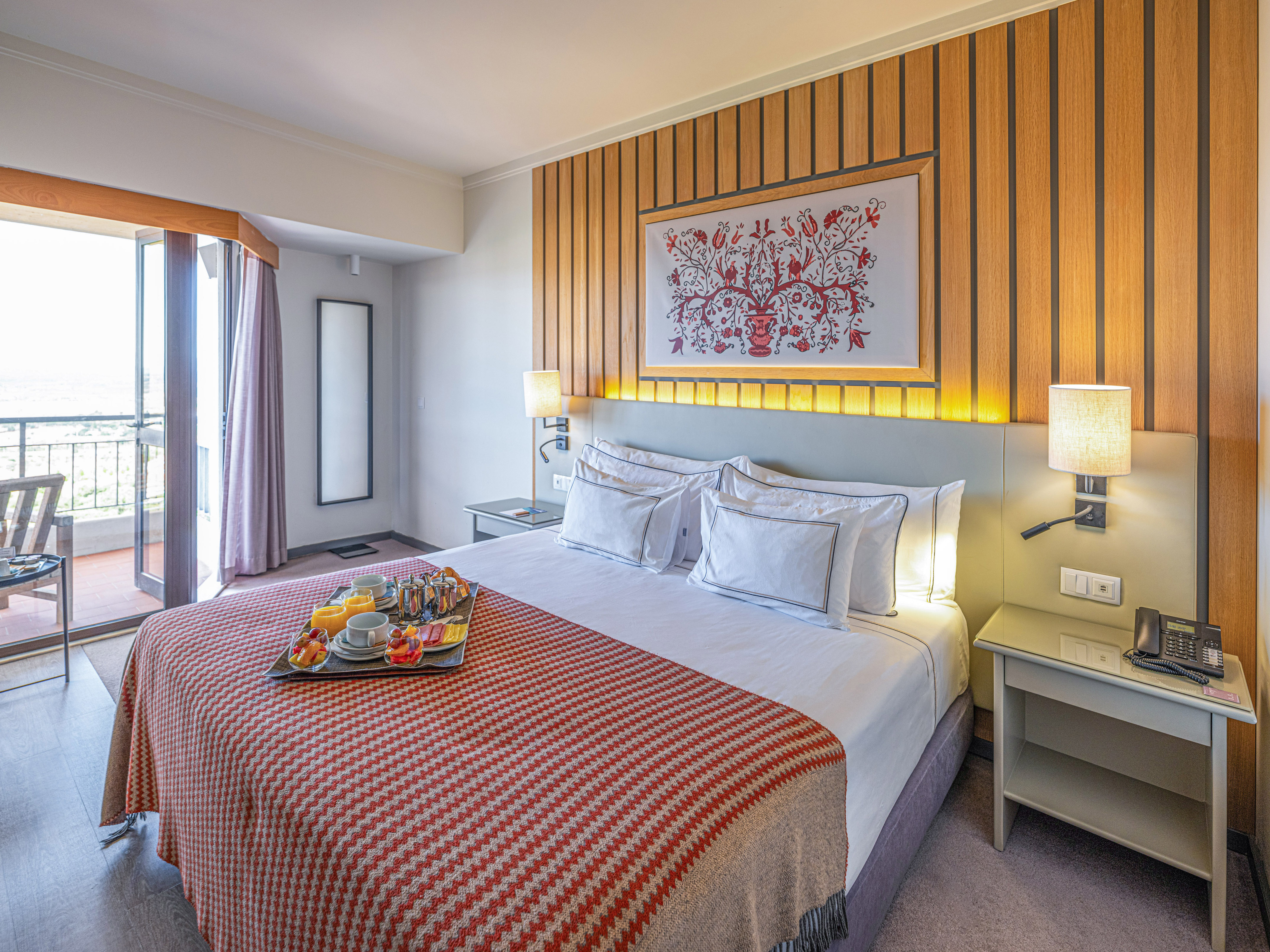 Hotel room with breakfast tray on bed, balcony, and wood wall decor.
