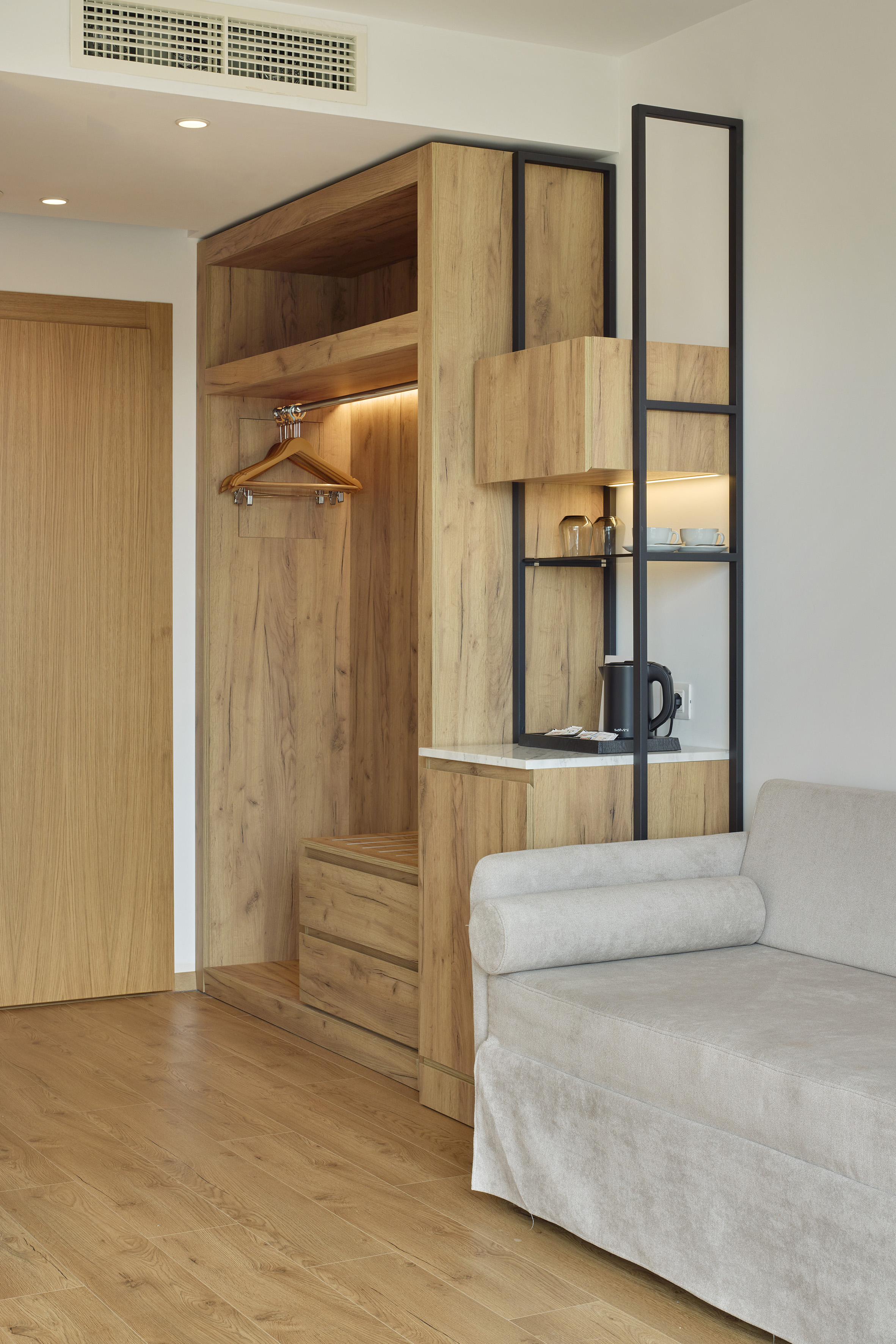 Modern hotel room with wooden wardrobe, integrated minibar, and light sofa.