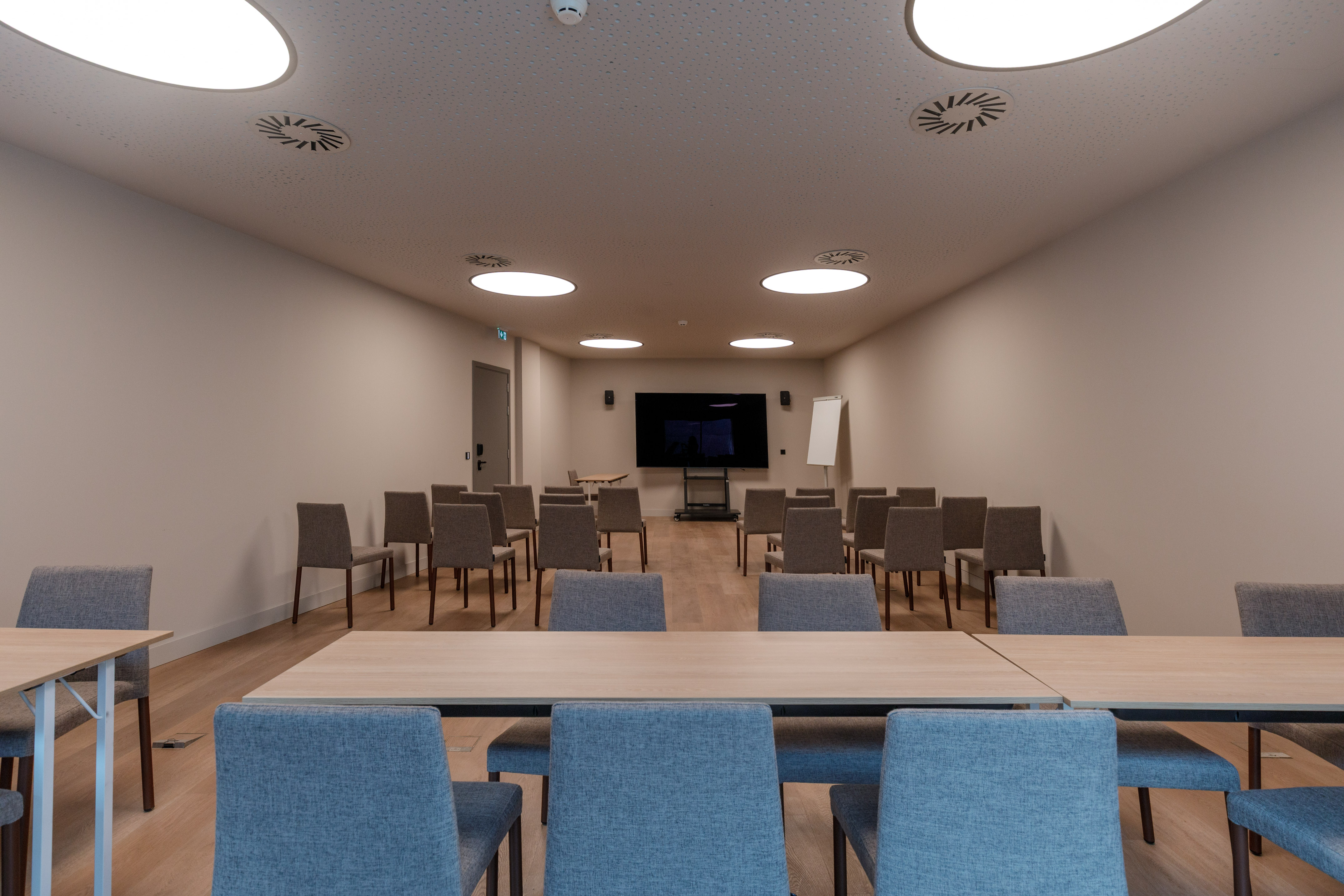 Modern empty meeting room with large screen, whiteboard, grey chairs, and light tables.