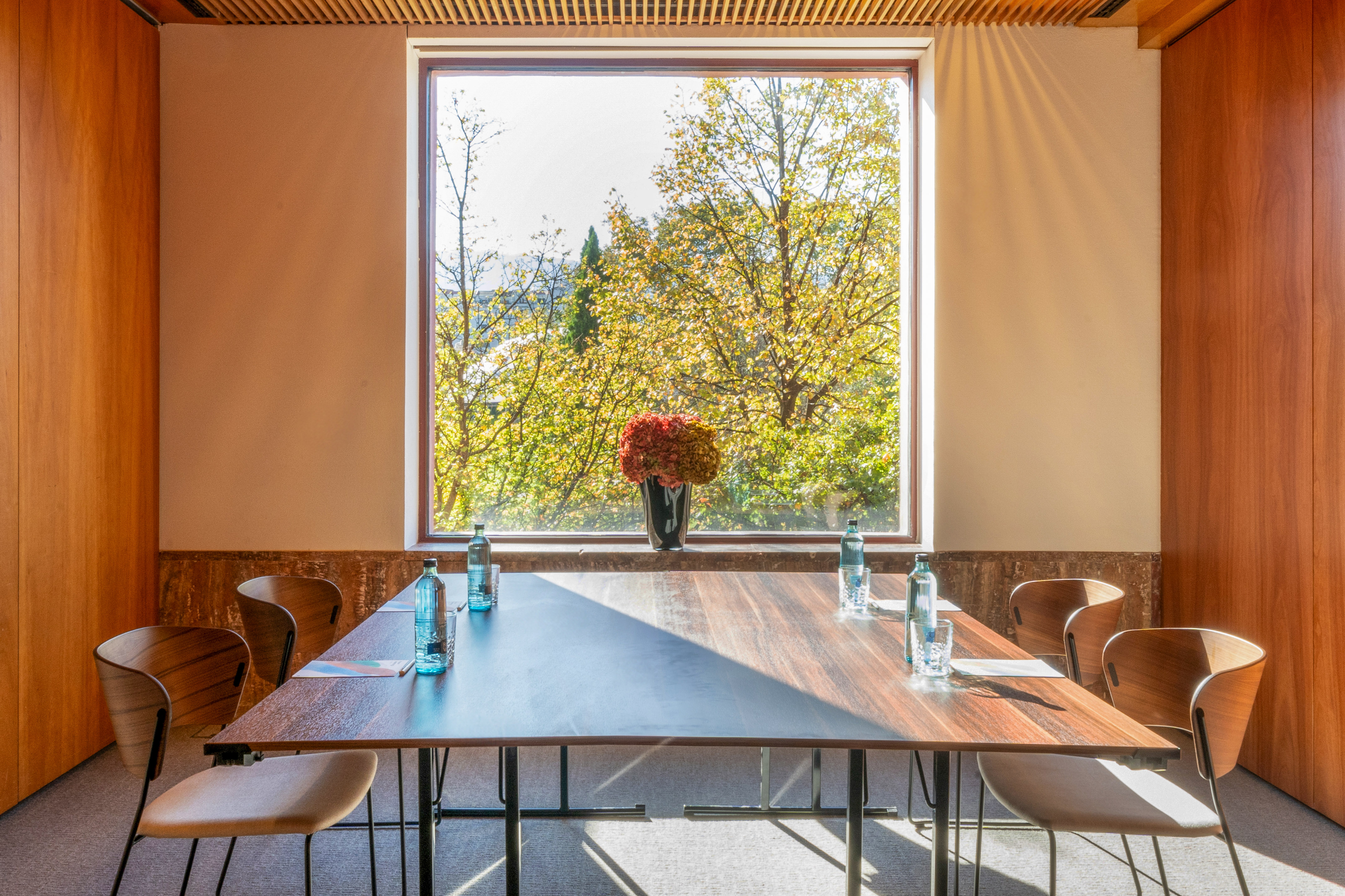 Modern meeting room, wooden table, chairs, large window with autumn trees.