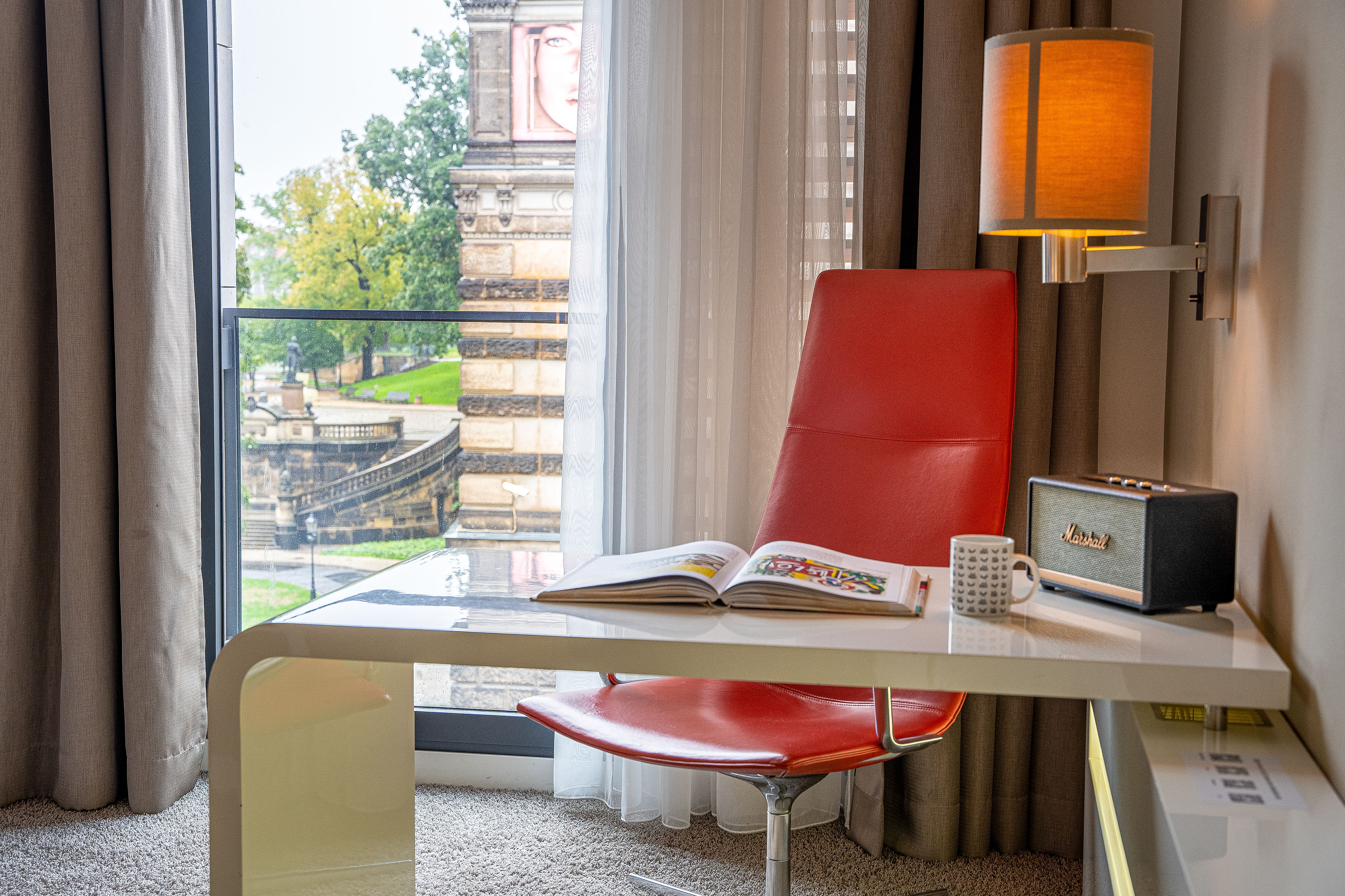 Modern hotel desk with red chair, Marshall speaker, and historic city view.