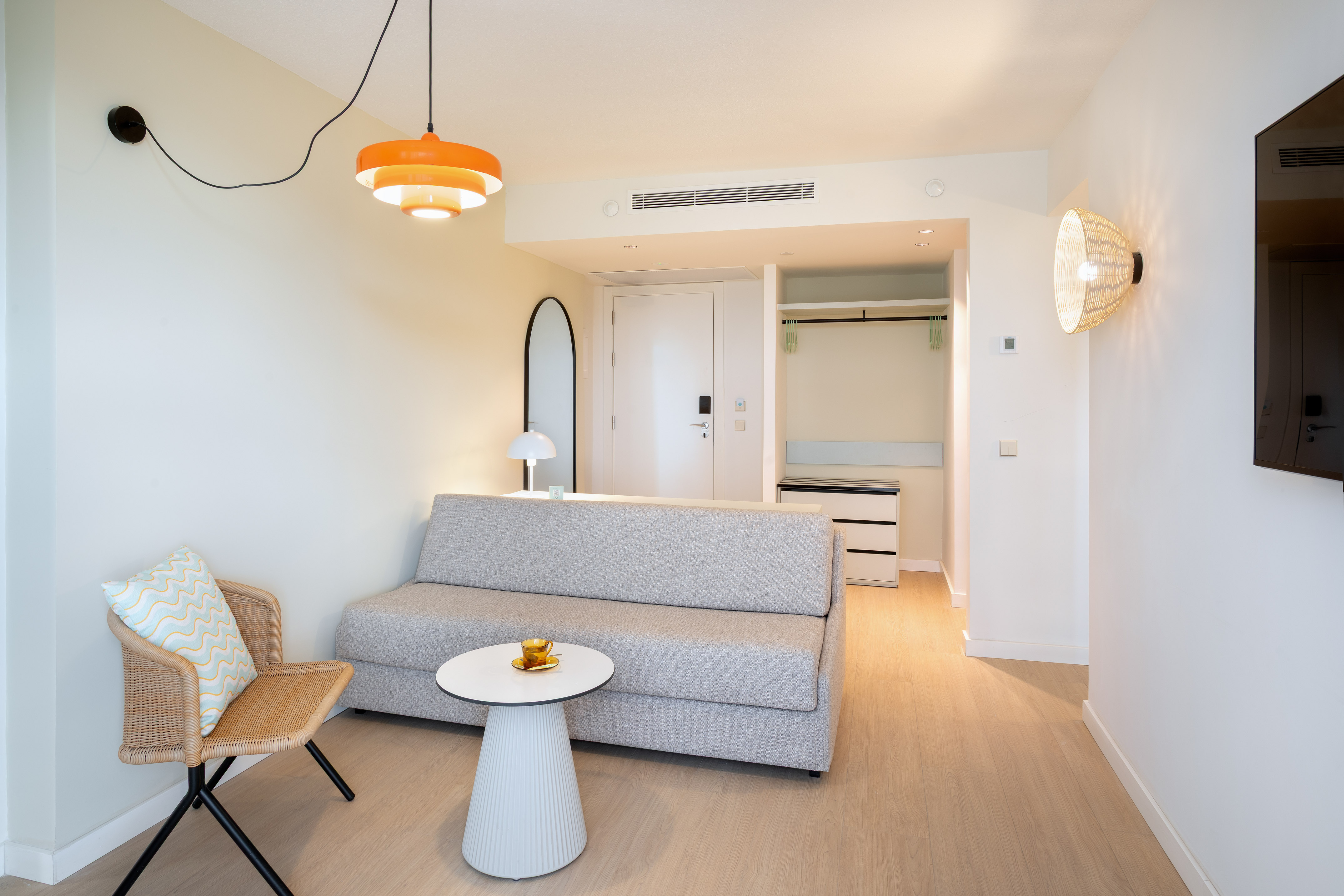 Modern hotel room with grey sofa bed, wicker chair, and orange pendant light.